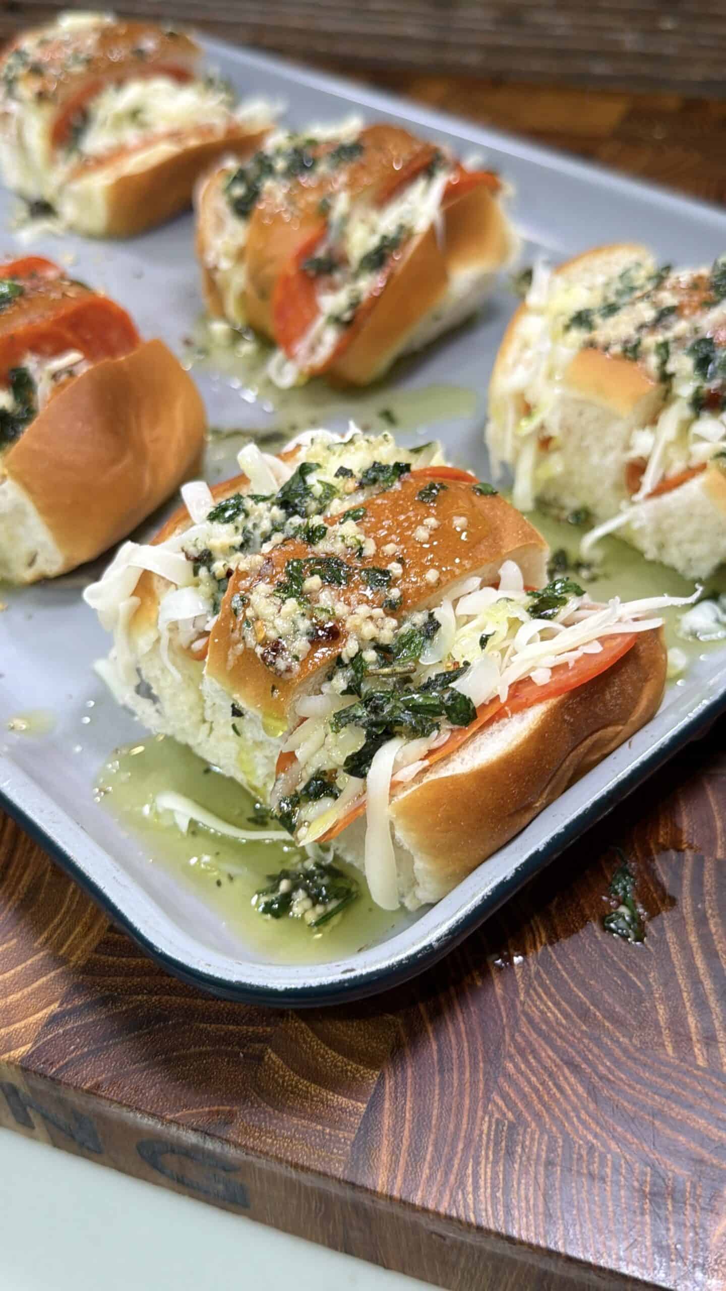 A tray of baked slider sandwiches filled with pepperoni, melted cheese, and topped with garlic herb butter, sitting on a wooden cutting board.