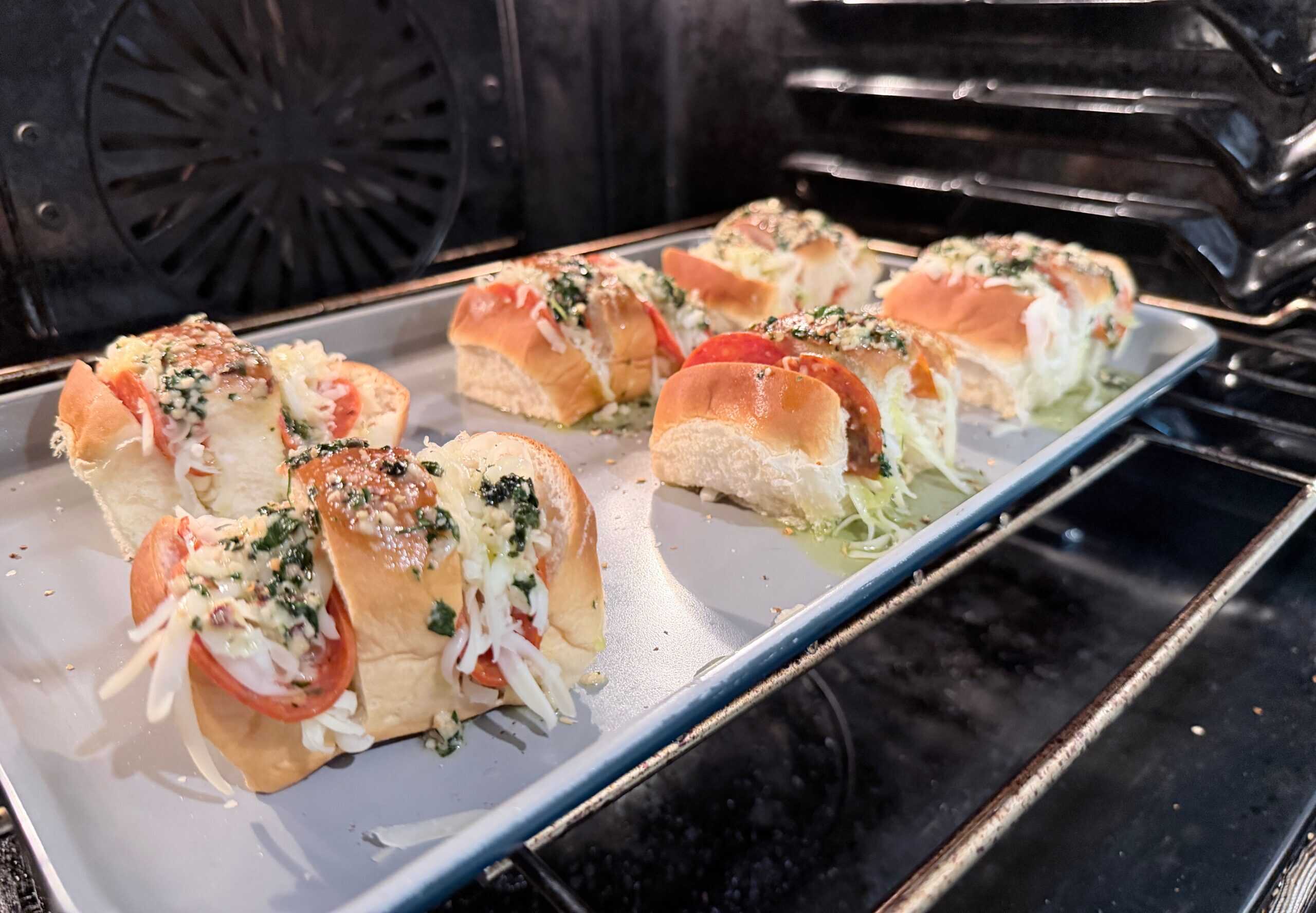 Six mini sandwiches with cheese, herbs, and tomato slices are arranged on a baking sheet inside an oven, ready to be baked. The bread is split and filled, with toppings spilling slightly onto the tray.