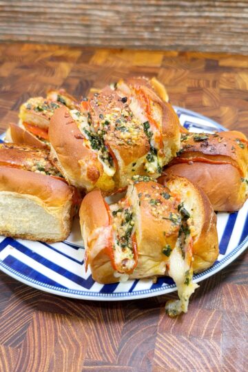 A plate with several cheese-stuffed bread rolls topped with herbs, melted cheese, and a drizzle of sauce, placed on a wooden surface.
