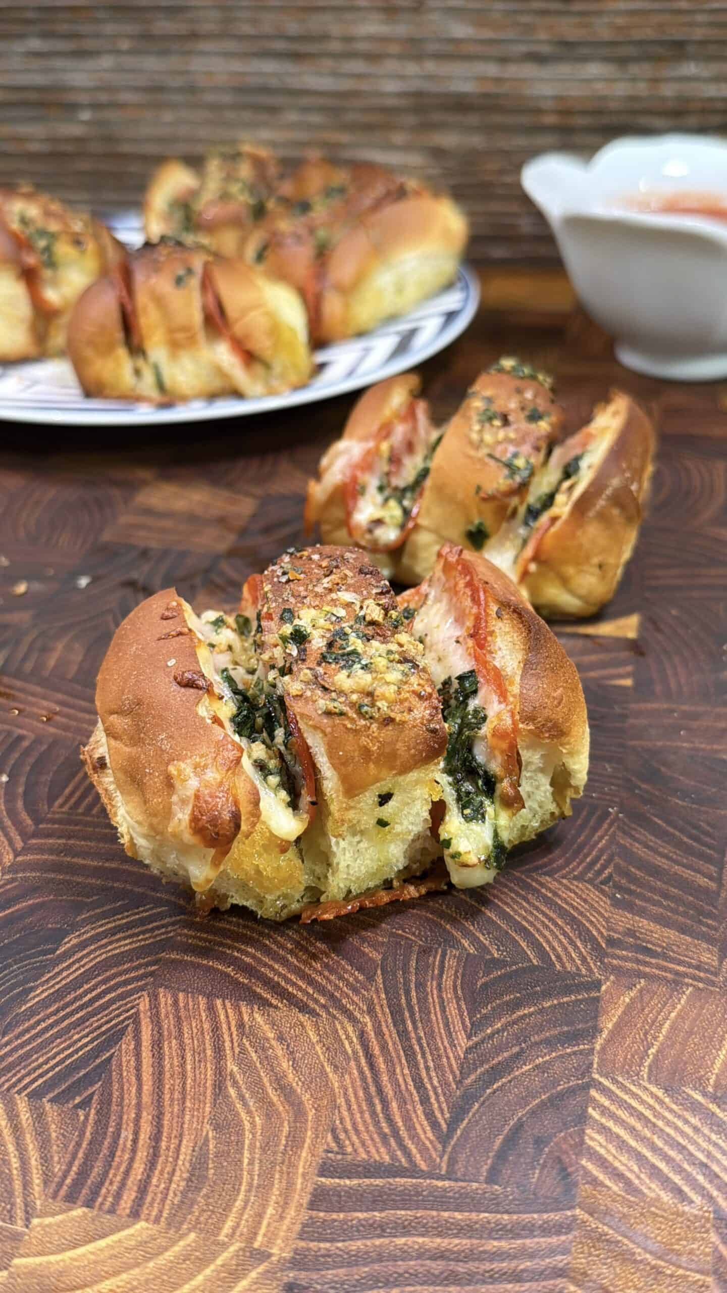 A close-up of a stuffed bread roll filled with melted cheese, spinach, and tomatoes, garnished with herbs, placed on a wooden surface. A plate with more rolls and a bowl of dipping sauce are in the background.