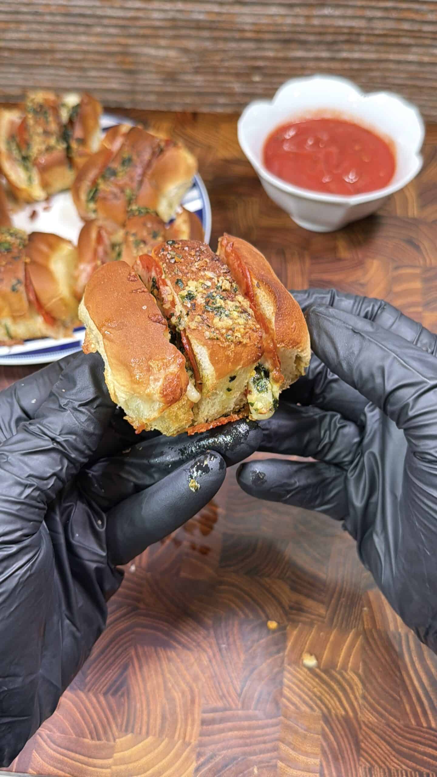 A person wearing black gloves holds a cheesy, stuffed slider pepperoni pizza pull apart. More sliders are on a plate in the background, with a bowl of red dipping sauce on a wooden surface nearby.