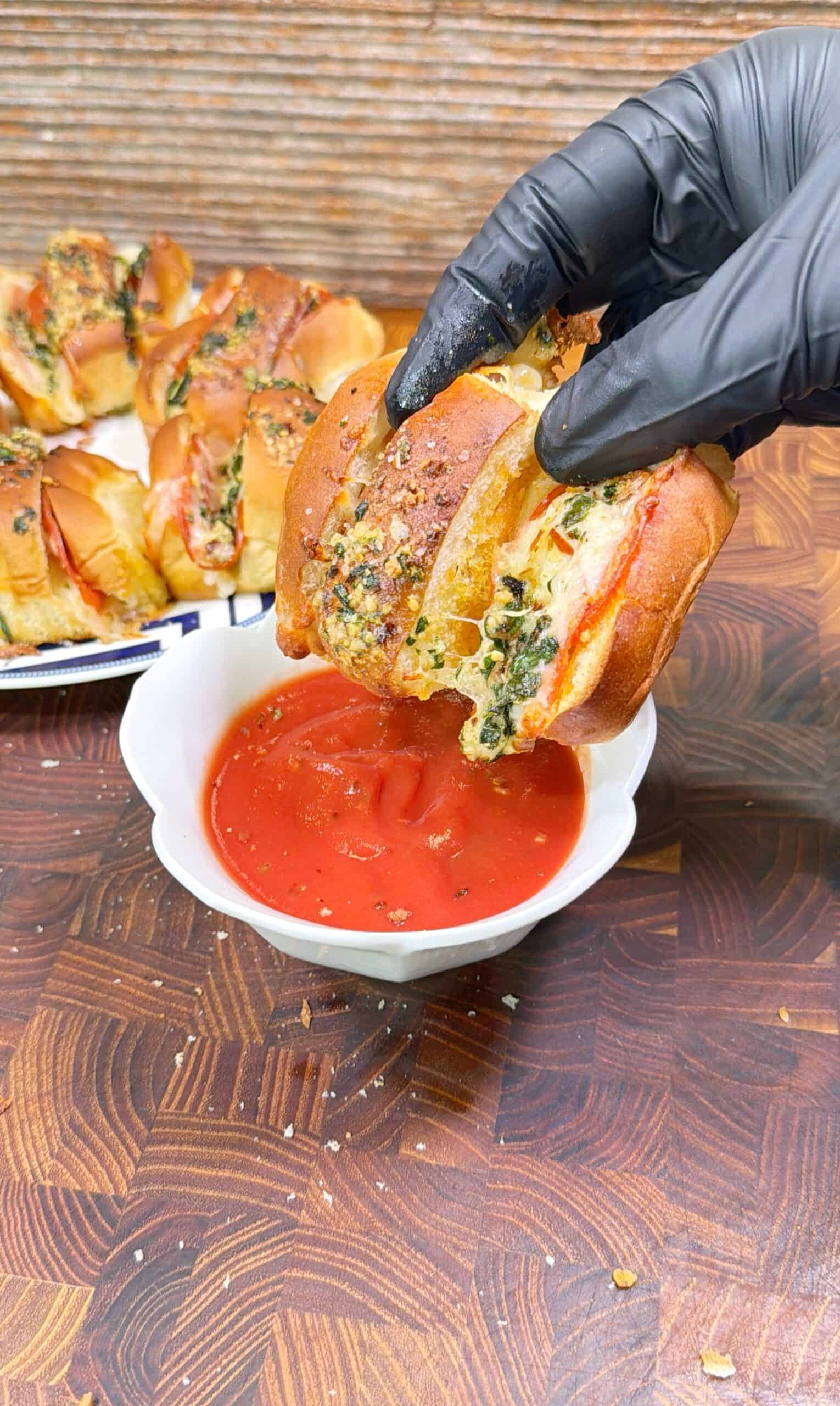 A gloved hand dips a stuffed bread slice filled with herbs and cheese into a bowl of tomato sauce. More stuffed bread slices are arranged on a plate in the background.