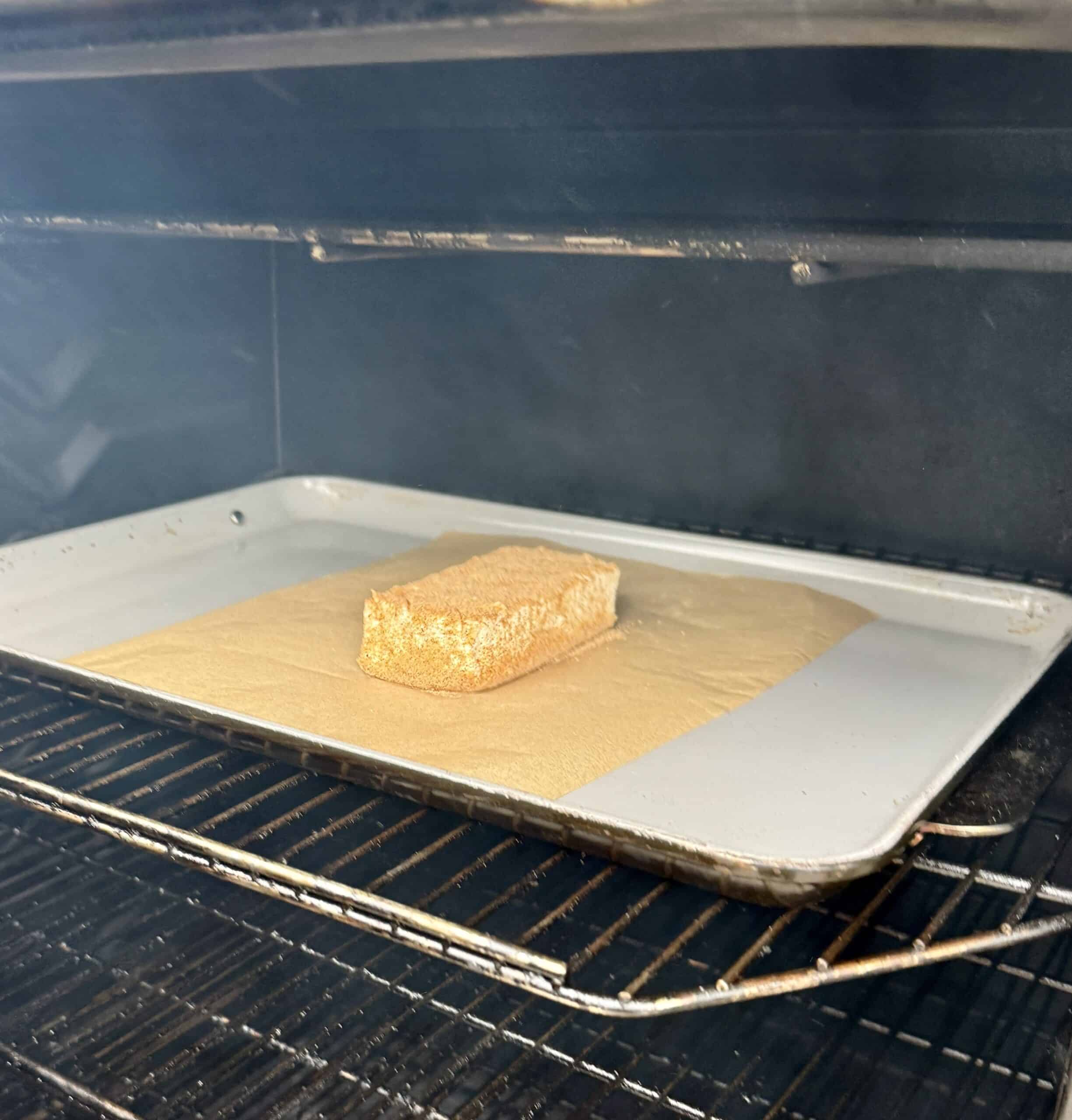 A single rectangular piece of breaded food, resembling smoked cream cheese, sits on parchment paper on a baking tray inside an oven. The oven racks and the open oven door are visible.