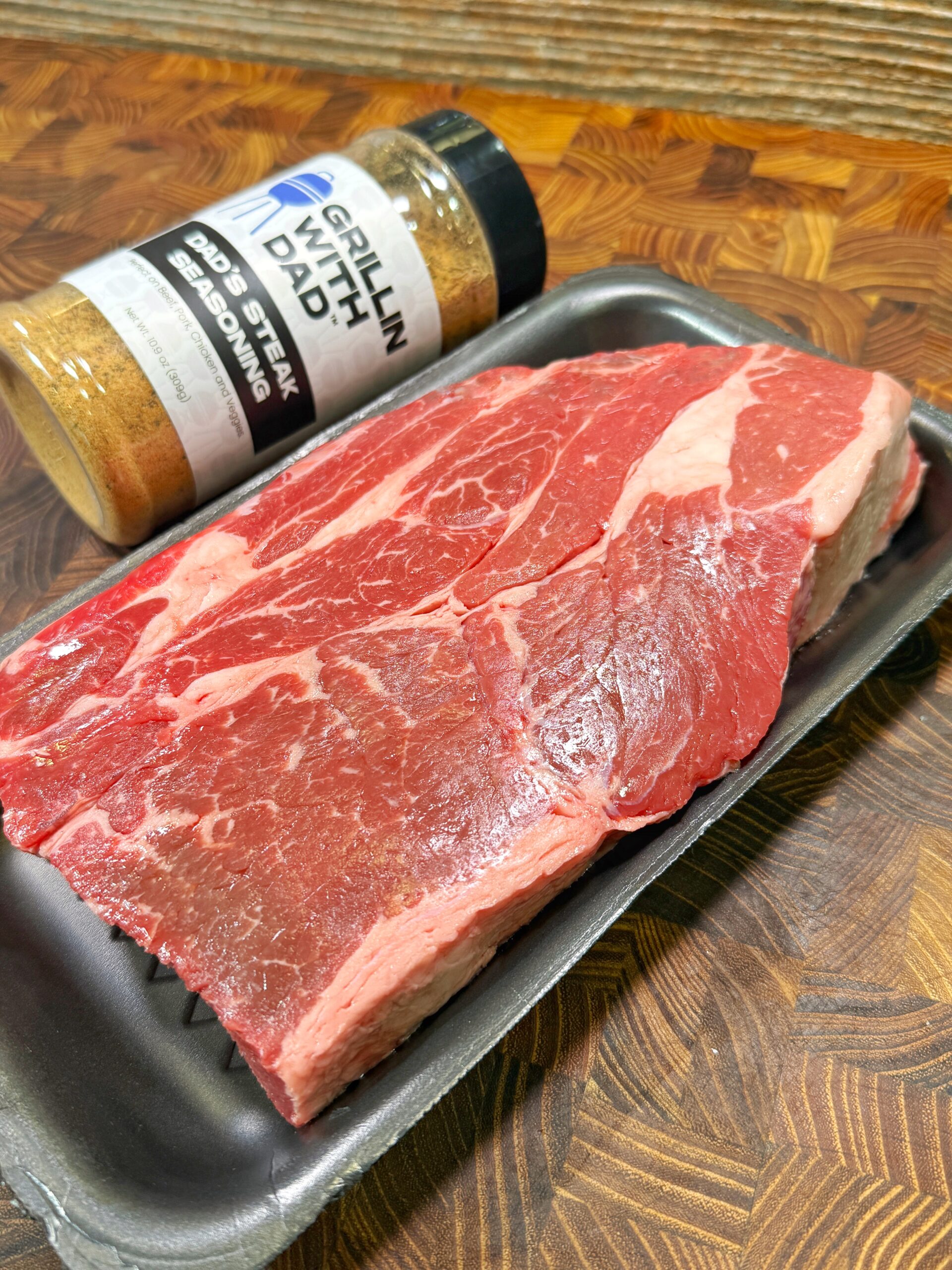 A raw steak sits in a black tray on a wooden surface, next to a container of Grillin’ With Dad 2nd Steak Seasoning.