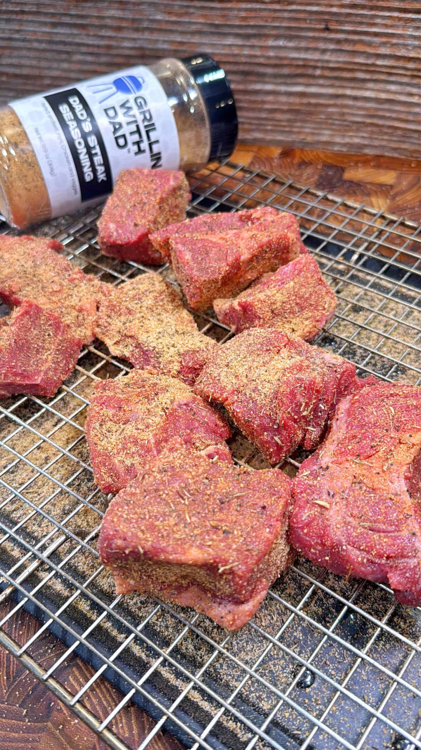 Several chunks of raw steak, coated with seasoning, rest on a wire rack. A seasoning bottle labeled “Grillin’ With Dad” lies open nearby, with its contents sprinkled on the steaks and rack.