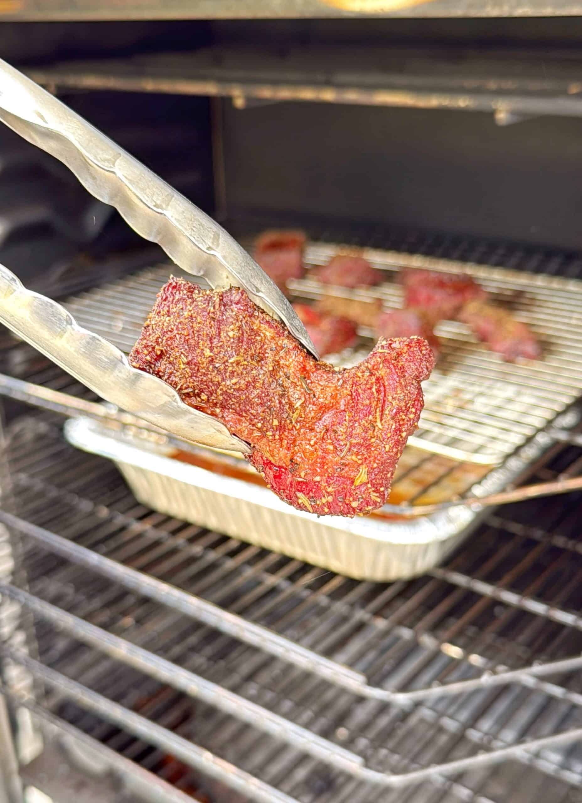 A close-up of metal tongs holding a piece of seasoned smoked meat inside a smoker, with more meat pieces on a wire rack above a foil pan in the background.