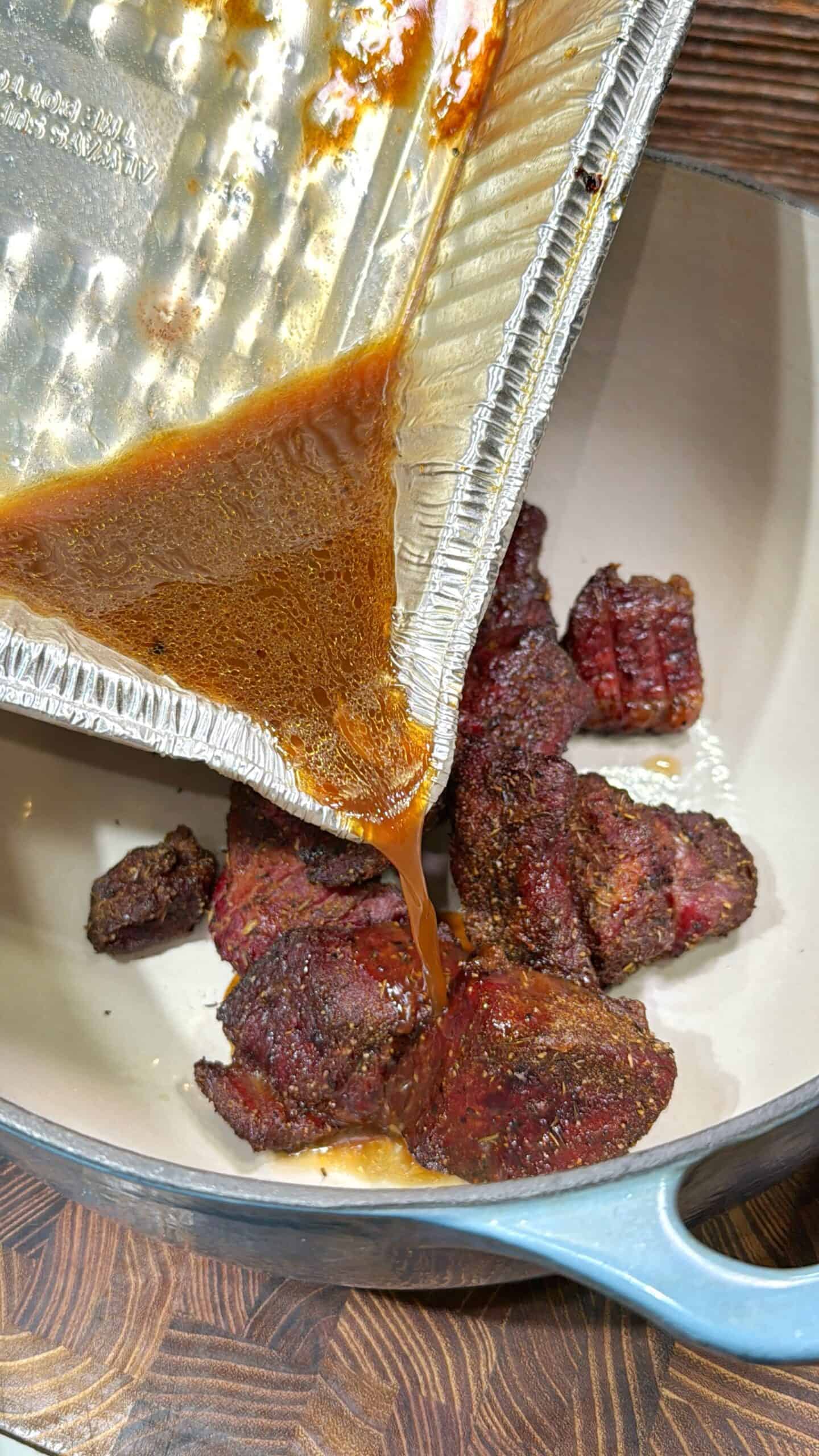 A close-up of cooked, seasoned meat chunks in a pot with sauce being poured over them from a foil tray. The pot sits on a wooden surface.