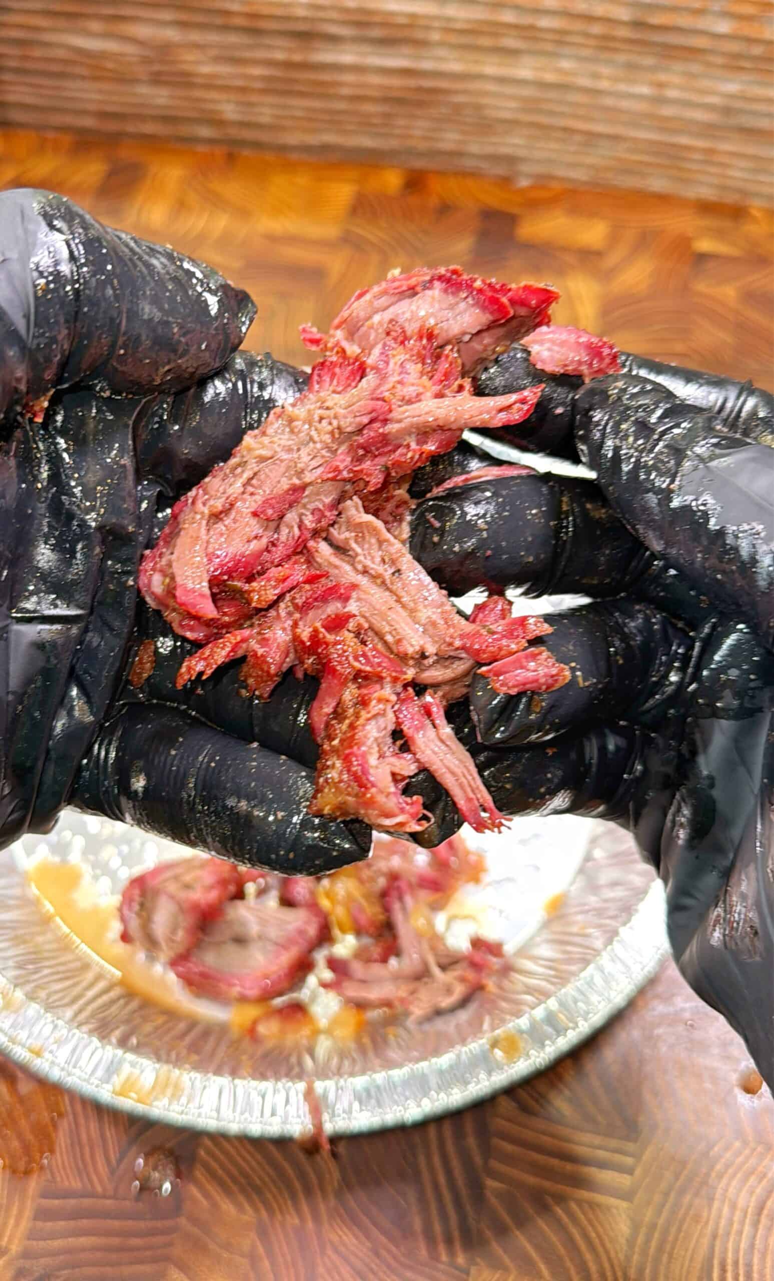 Gloved hands pull apart juicy, smoked meat over a foil plate on a wooden surface, showing tender, shredded texture and reddish color.