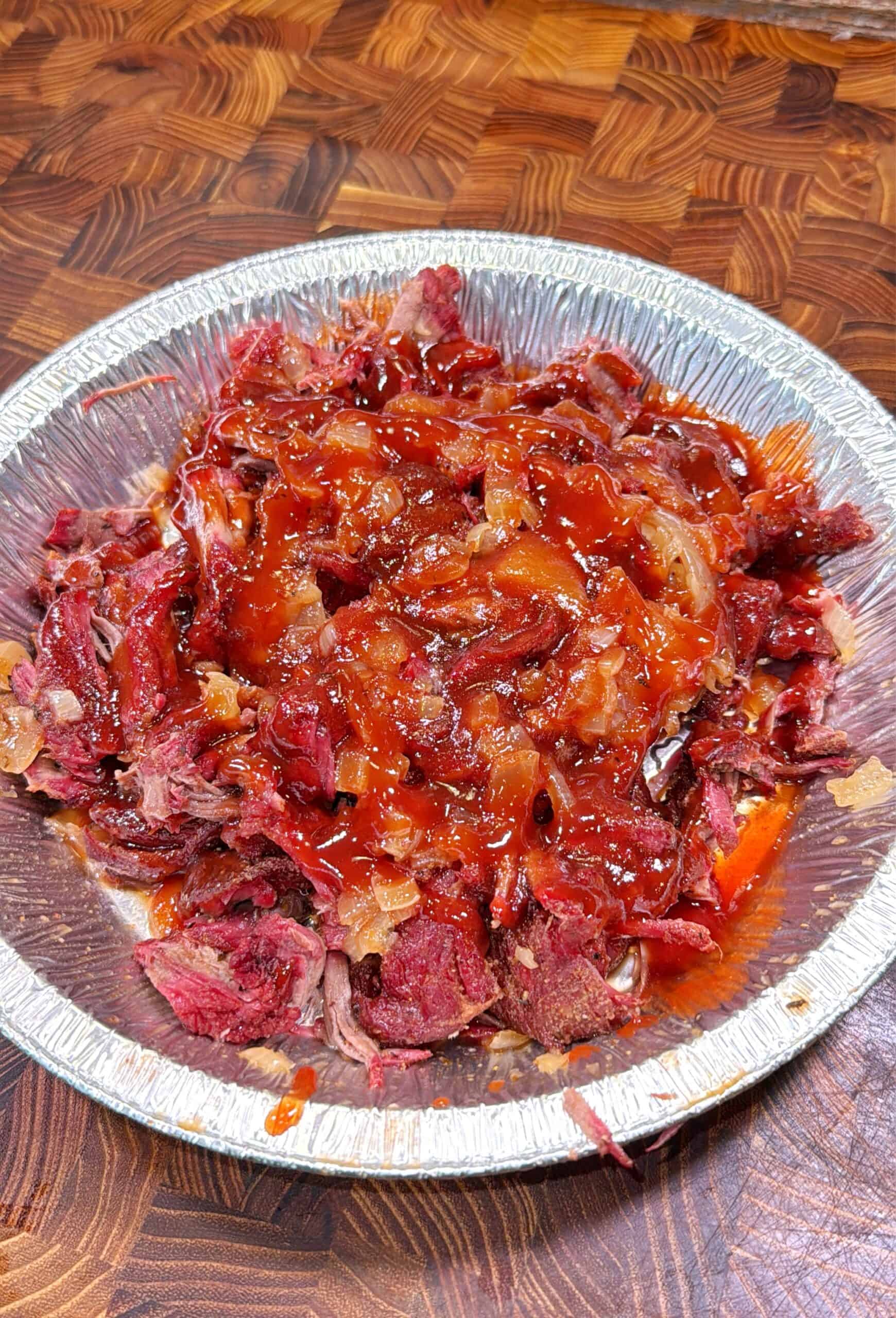 A round aluminum container filled with shredded barbecue beef covered in a layer of reddish-brown barbecue sauce, set on a wooden cutting board.
