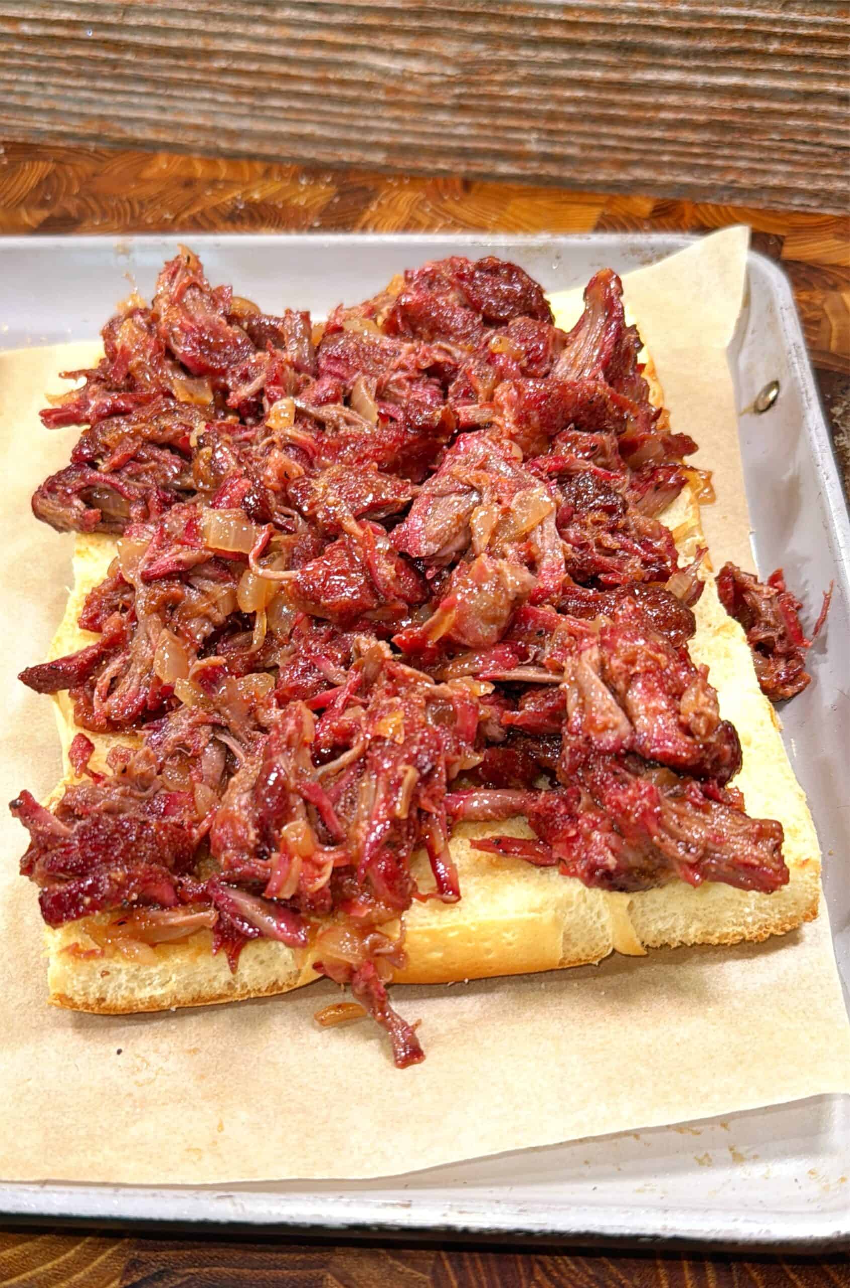 Pulled beef with caramelized onions piled on a sliced, toasted hoagie roll, served on parchment paper over a metal tray, with a rustic wooden background.
