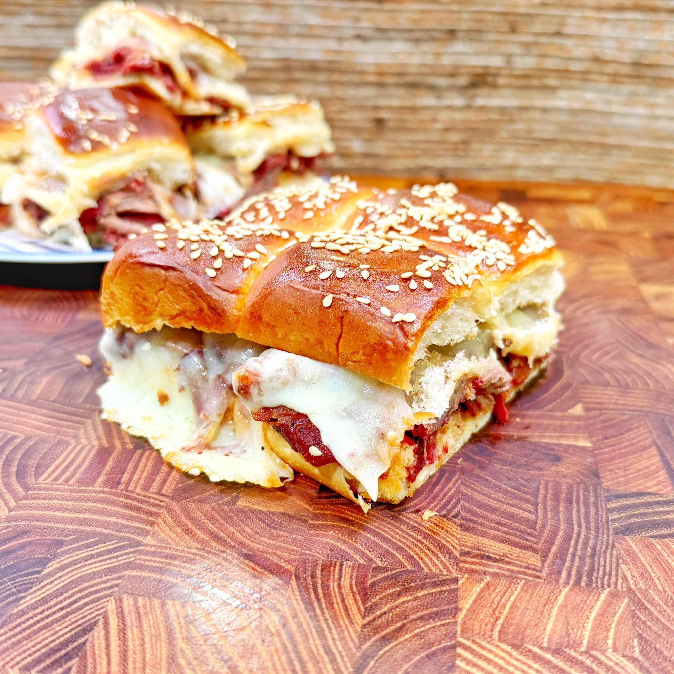 A close-up of a bbq beef sliders sandwich with sesame-topped bread, filled with melted cheese and deli meat, resting on a wooden surface; more sandwiches are stacked on a plate in the background.