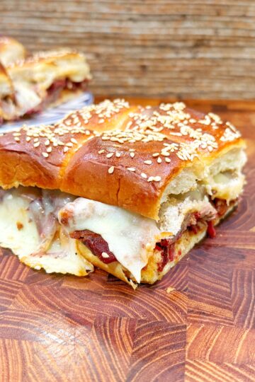 A close-up of a sesame-topped sandwich filled with melted cheese and deli meat, resting on a wooden cutting board. In the background, more sandwich pieces sit on a plate.