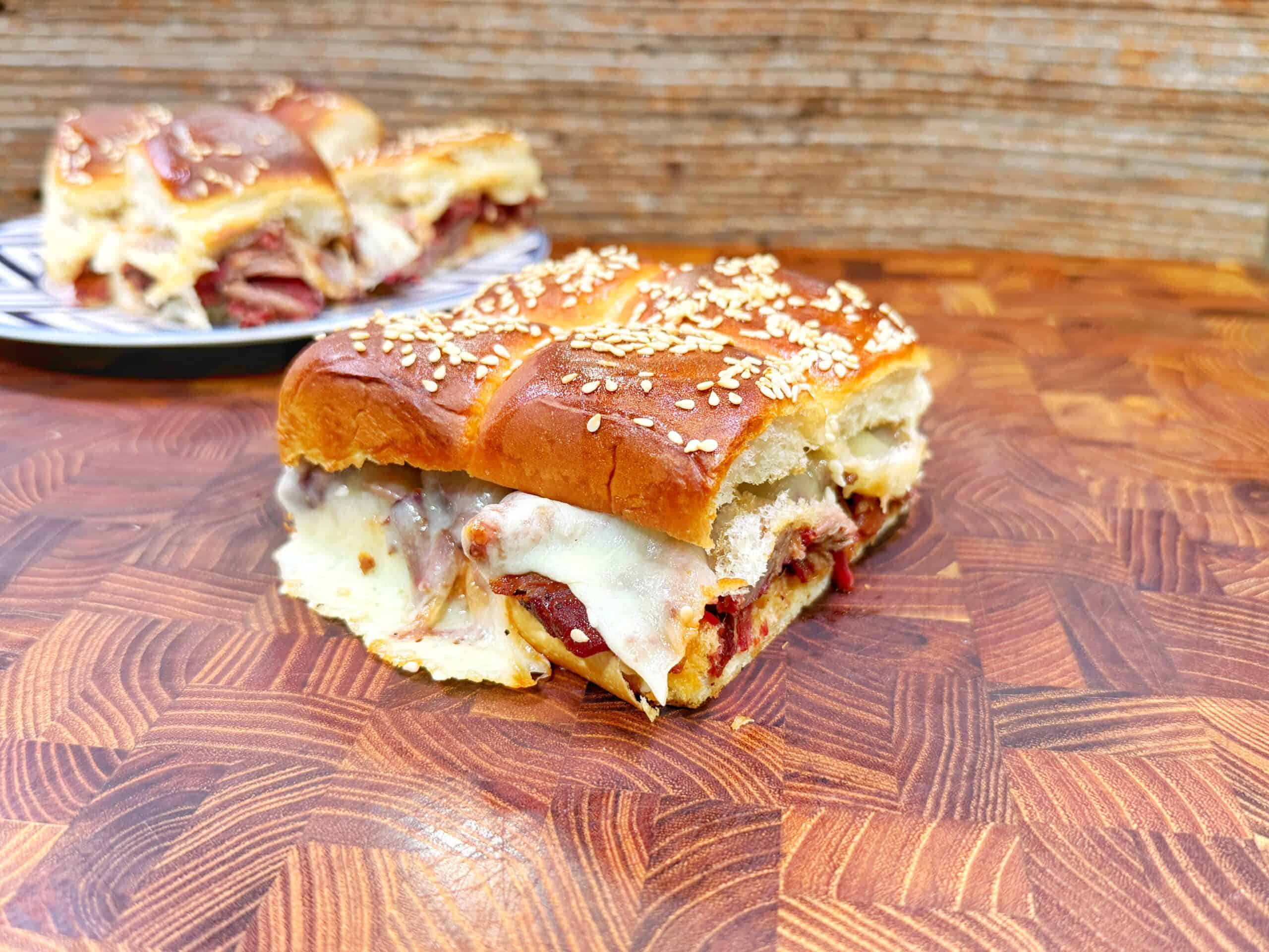 A close-up of a sesame-topped sandwich filled with melted cheese and deli meat, resting on a wooden cutting board. In the background, more sandwich pieces sit on a plate.