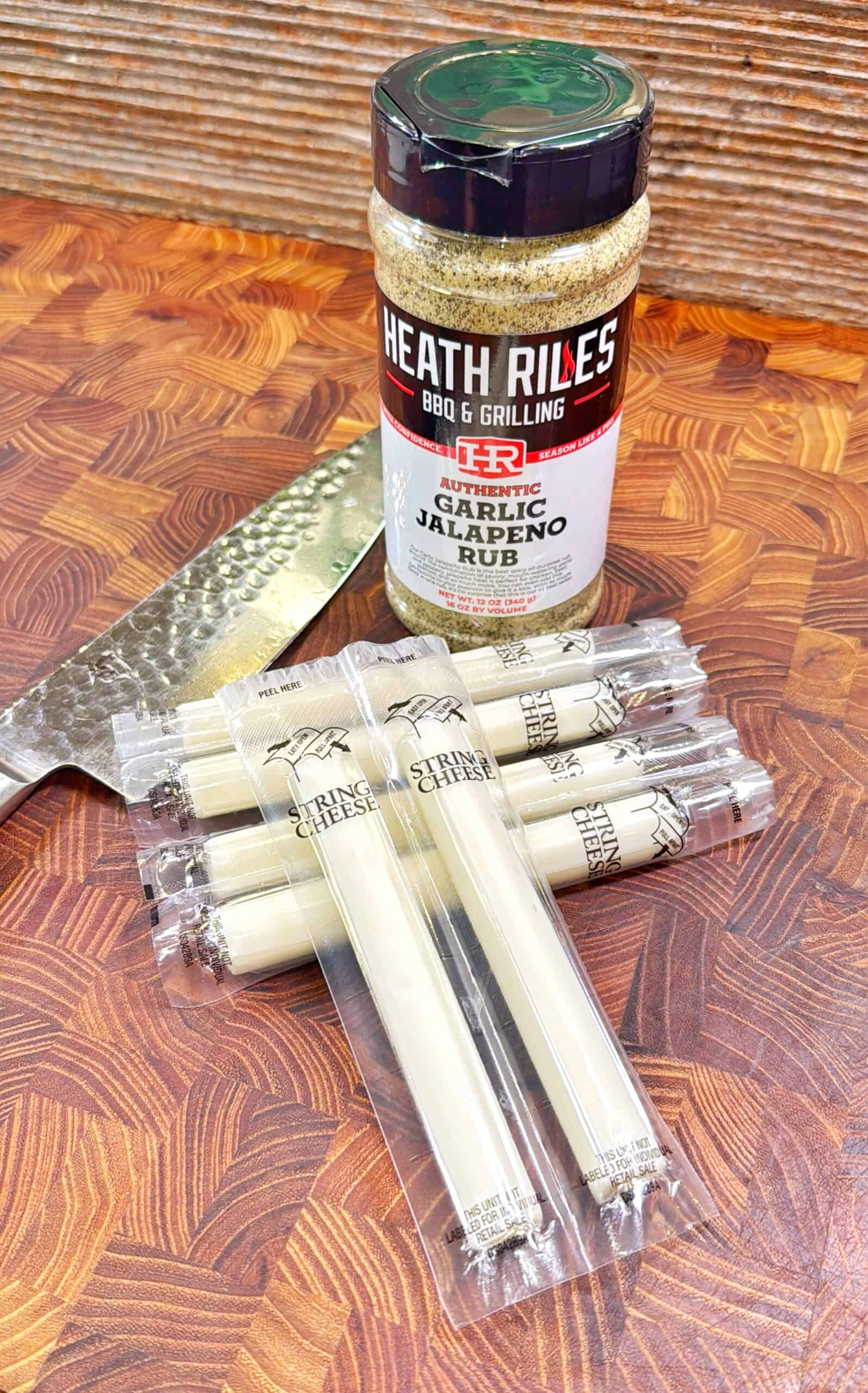 A jar of Heath Riles Garlic Jalapeno Rub, a chef’s knife, and seven individually wrapped string cheese sticks are arranged on a wooden cutting board.