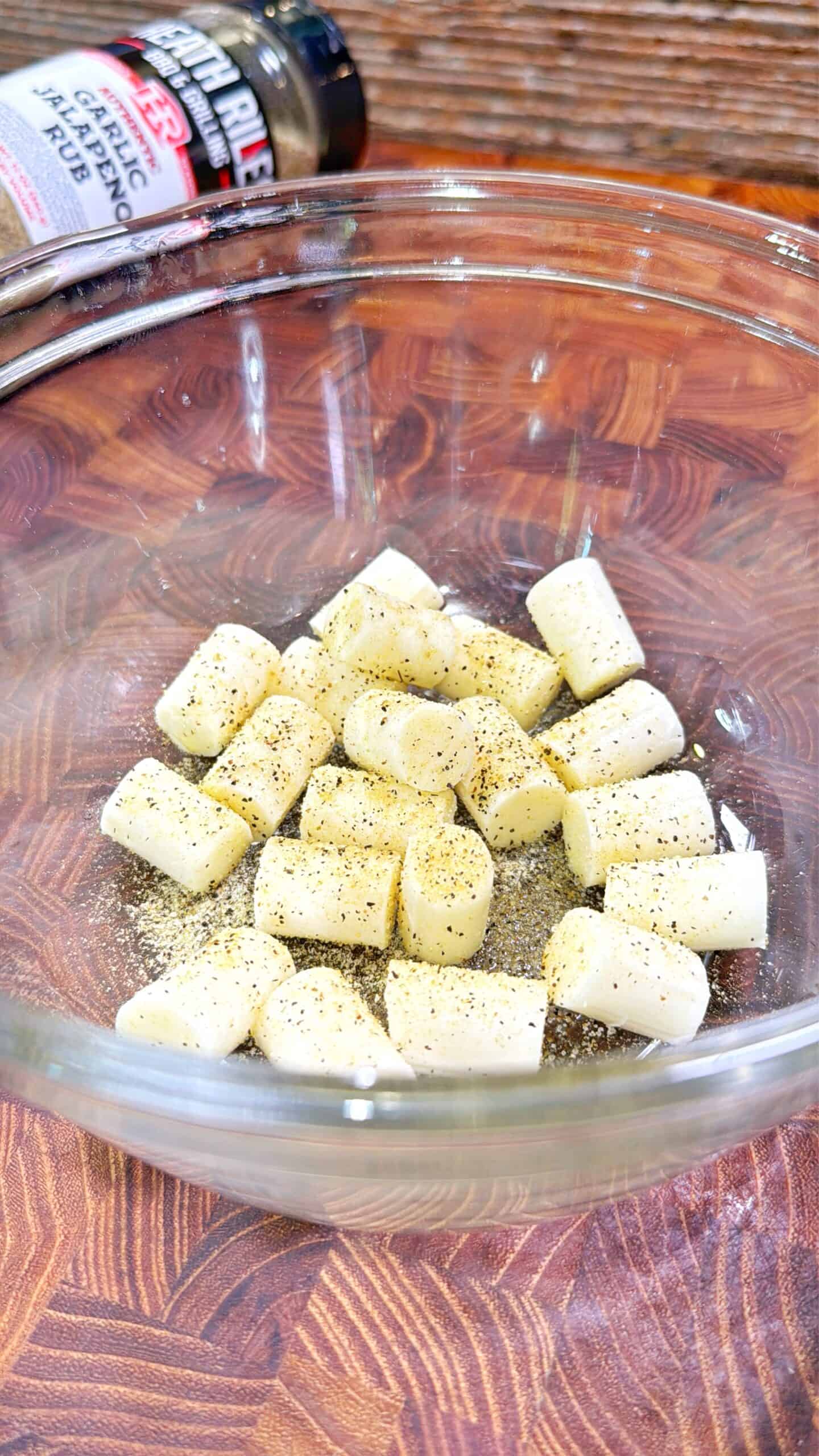 A glass bowl filled with chunks of garlic topped with black pepper sits on a wooden cutting board. A container of garlic jalapeño rub is partially visible in the background.