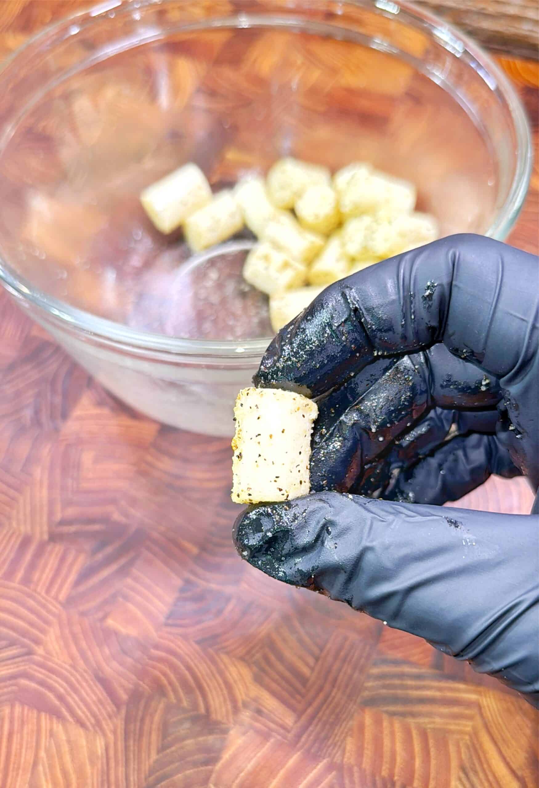 A gloved hand holds a seasoned gnocchi piece above a glass bowl containing more gnocchi pieces, set on a wooden surface.