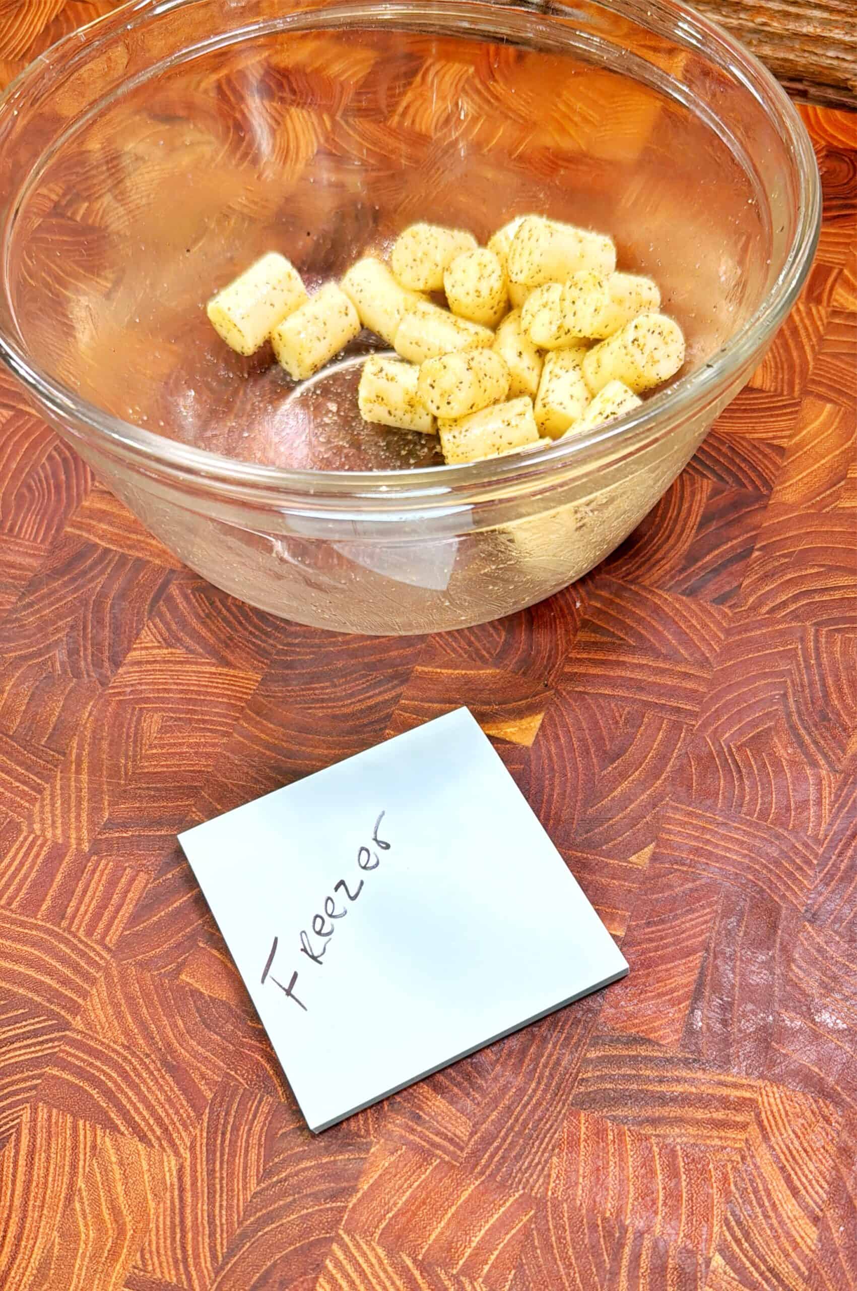 A glass bowl with chopped bananas sprinkled with cinnamon sits on a patterned wooden surface. In front of the bowl is a handwritten sticky note labeled Freezer.