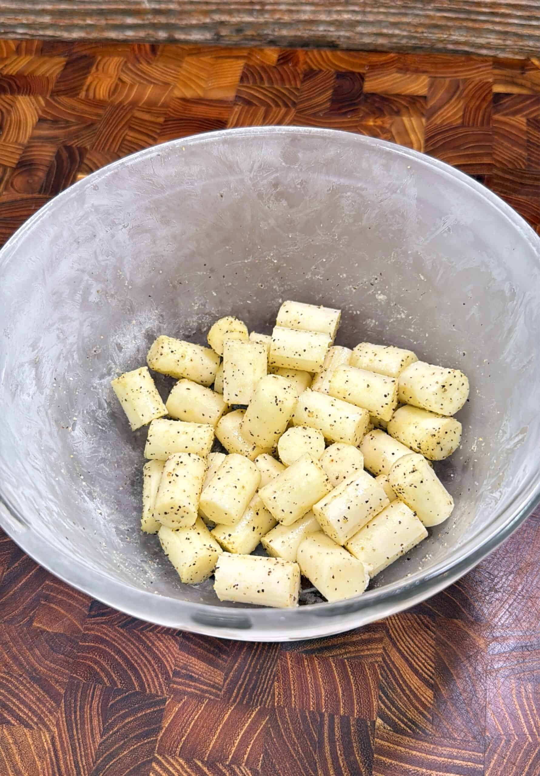 A glass bowl filled with cubed gnocchi coated in oil, black pepper, and seasoning, sitting on a wooden cutting board surface.