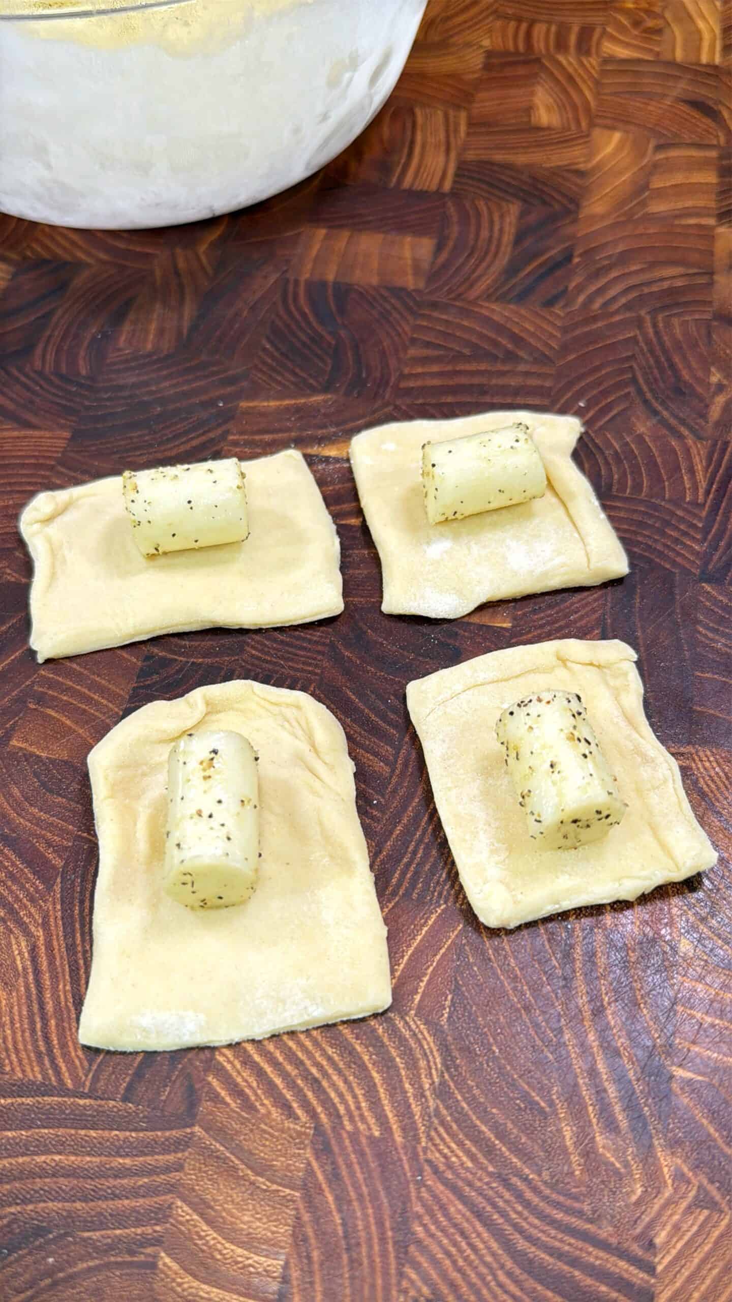 Four pieces of pasta dough squares each topped with a small, seasoned cylinder of cheese, are arranged on a wooden cutting board. A bowl is partially visible in the top left corner.