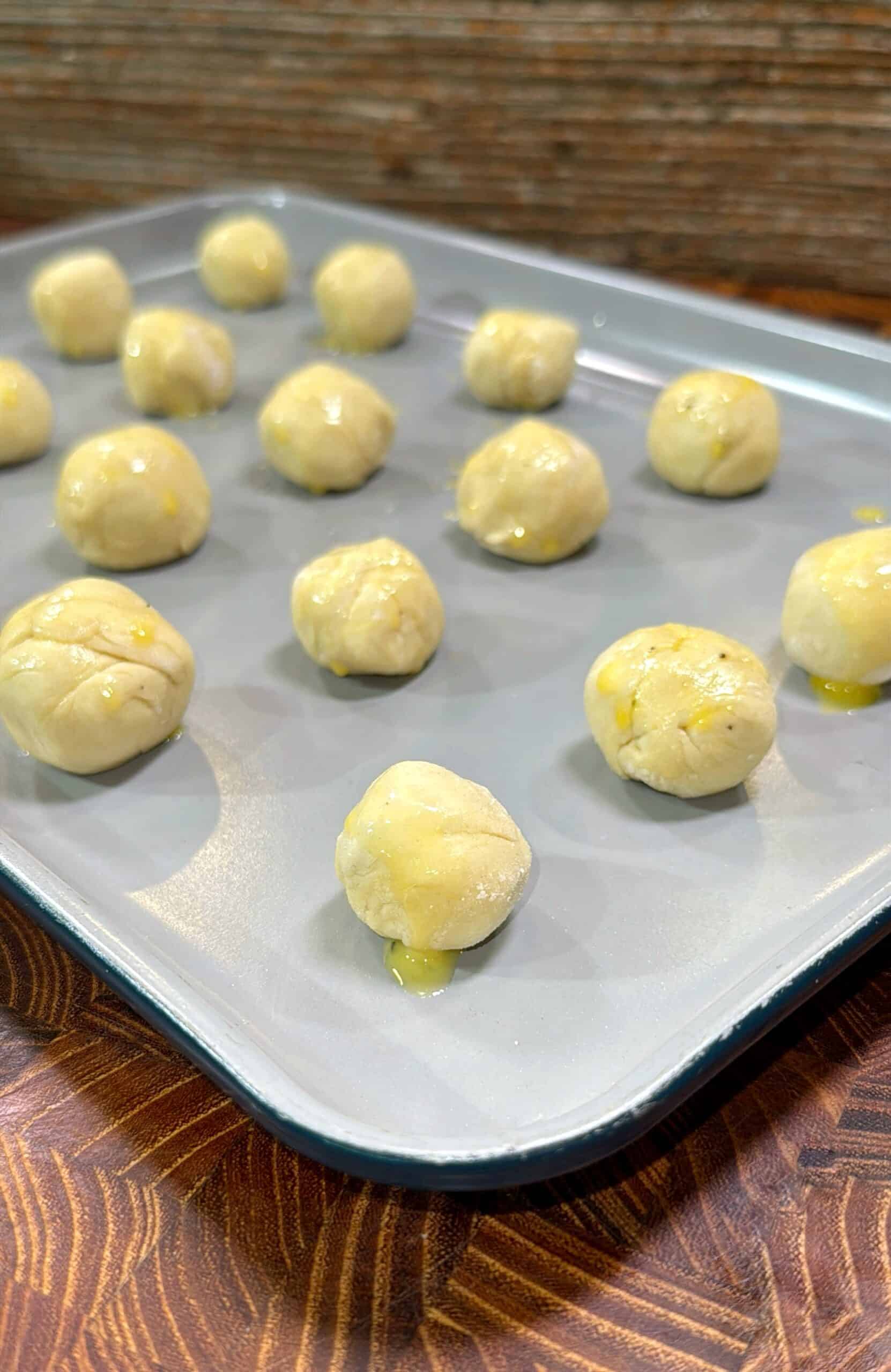 Unbaked dough balls, lightly brushed with oil, are arranged on a gray baking sheet, ready to be baked. The background features a wooden surface and a textured brown backdrop.