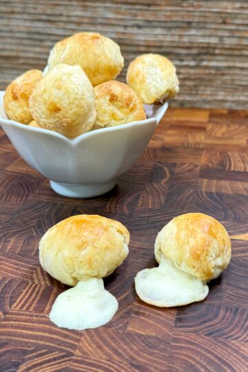 A white bowl filled with round, golden cheese puffs sits on a wooden surface. Two cheese puffs in front of the bowl have melted cheese oozing out. The background features a rustic wooden wall.