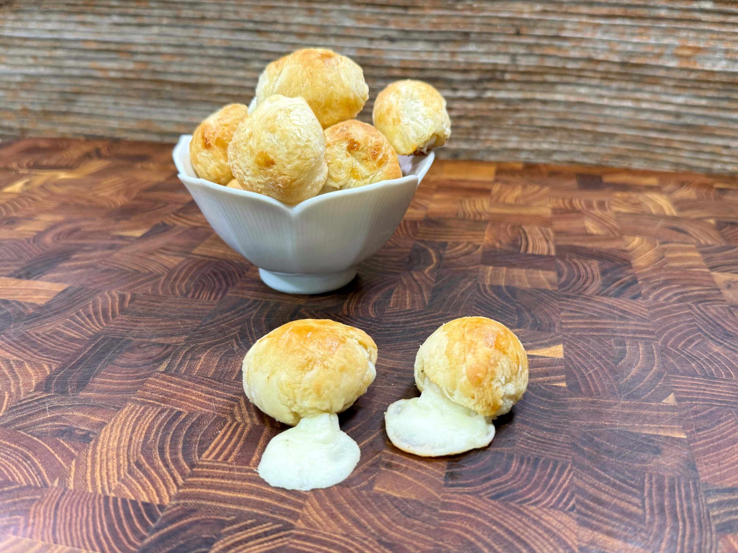 A white bowl filled with round, golden cheesy bread balls sits on a wooden surface. Two cheese puffs in front of the bowl have melted cheese oozing out. The background features a rustic wooden wall.