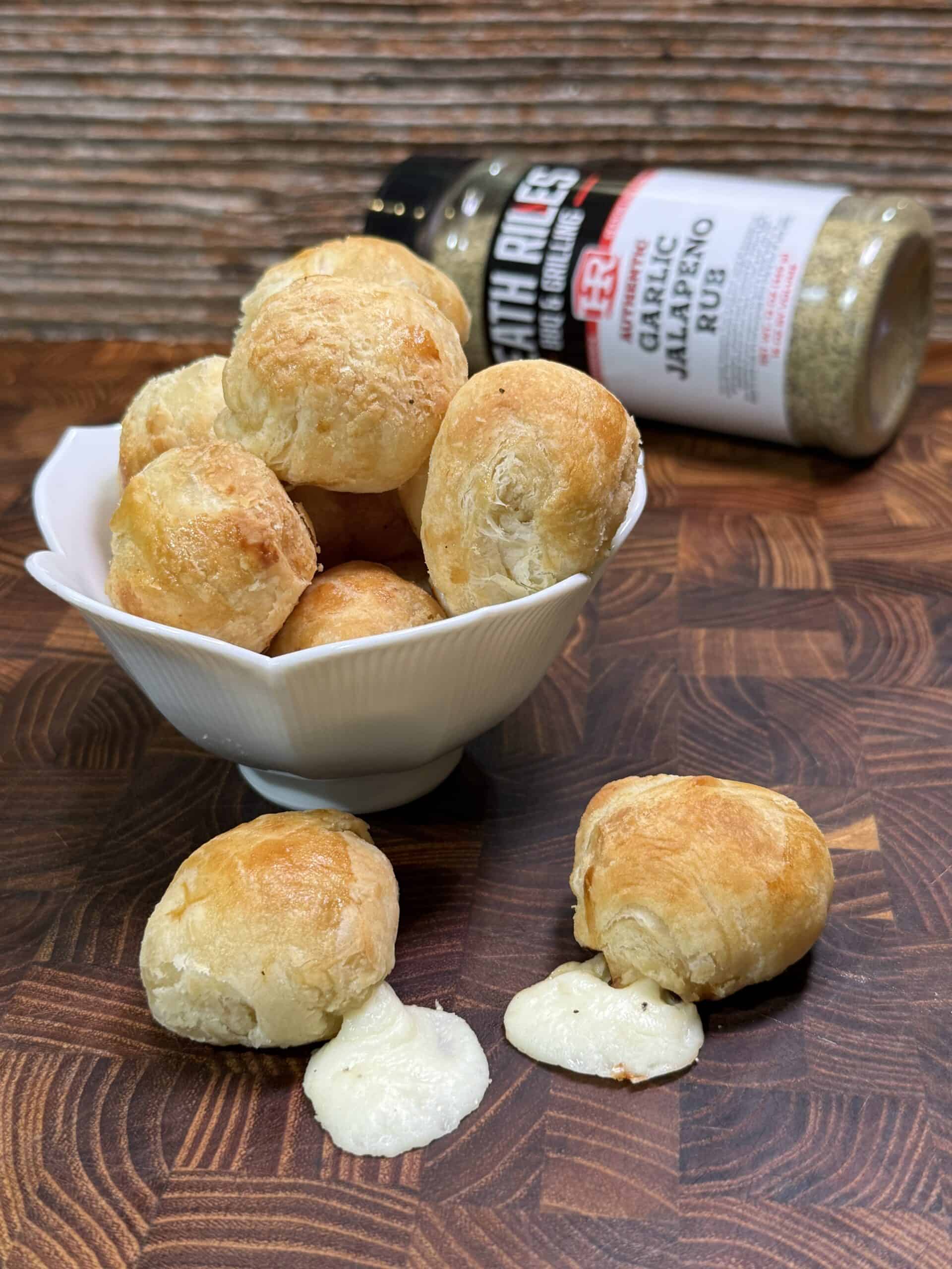 A bowl of golden-brown cheese-stuffed rolls sits on a wooden surface, with two rolls in front, one oozing melted cheese. A seasoning bottle labeled Garlic Jalapeno Rub lies in the background.