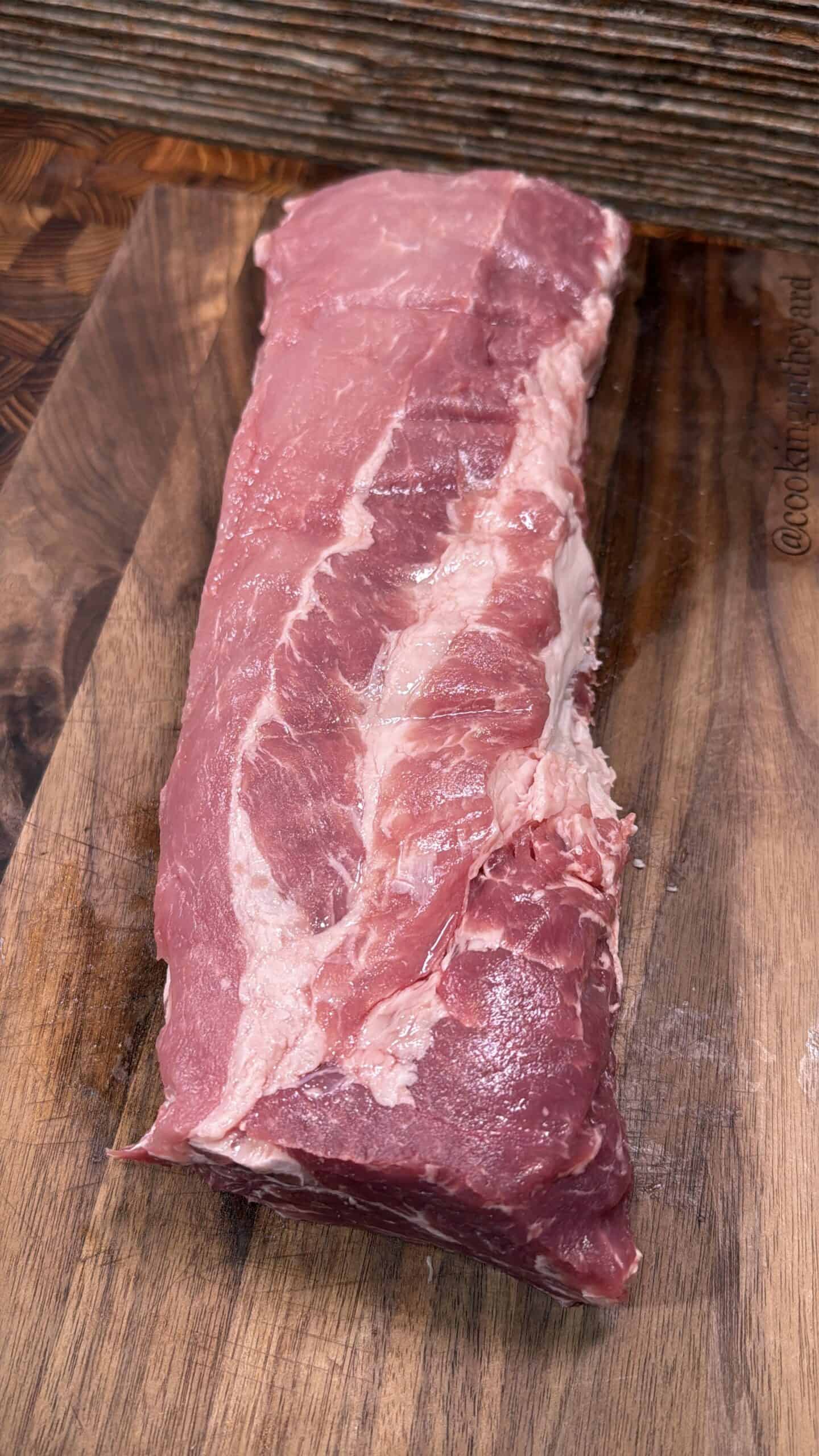 A raw pork loin with visible fat marbling is placed on a wooden cutting board, ready for preparation.