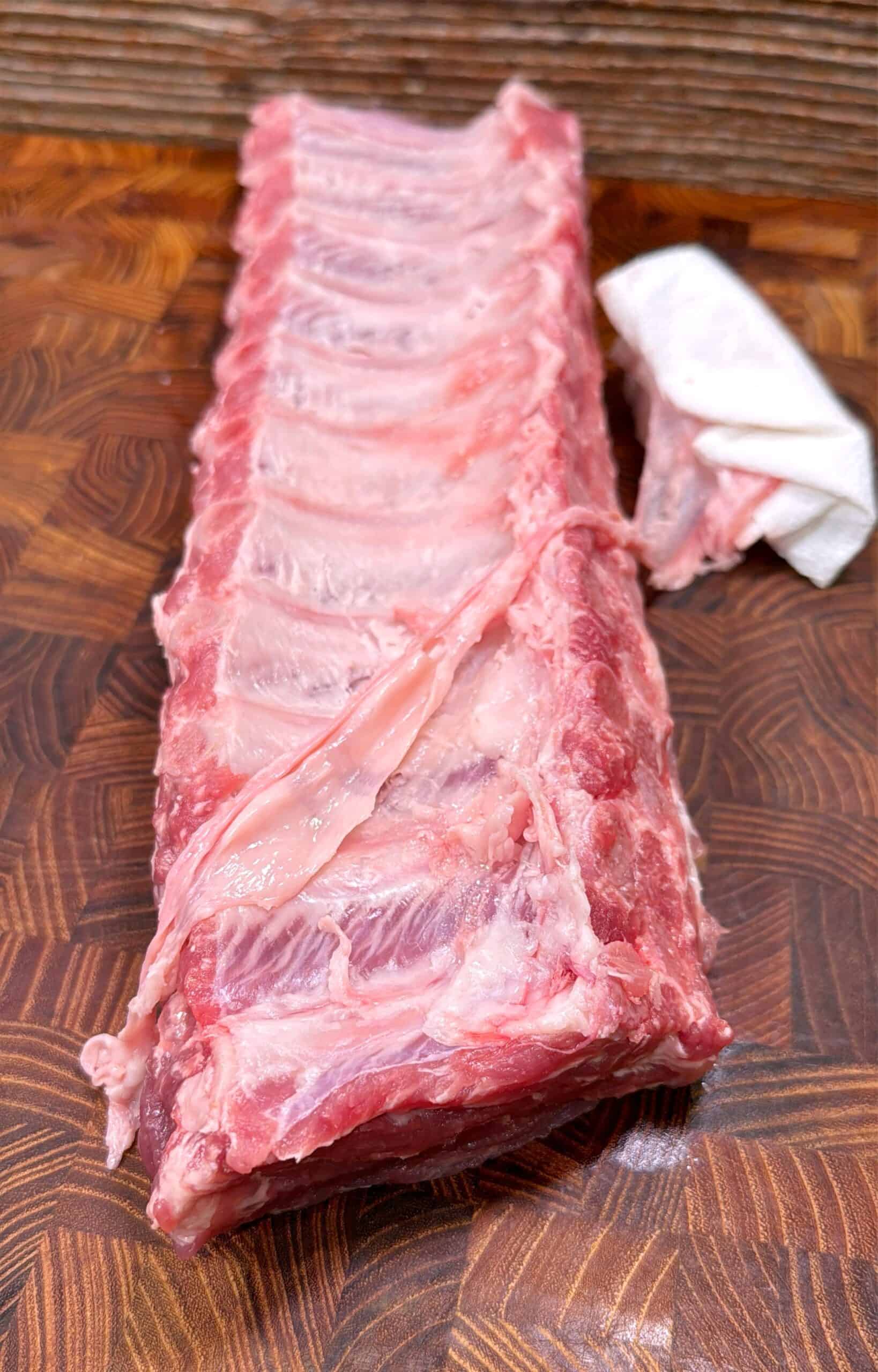 A rack of raw pork ribs on a wooden cutting board, with a paper towel gripping the thin membrane being peeled from the back of the ribs.