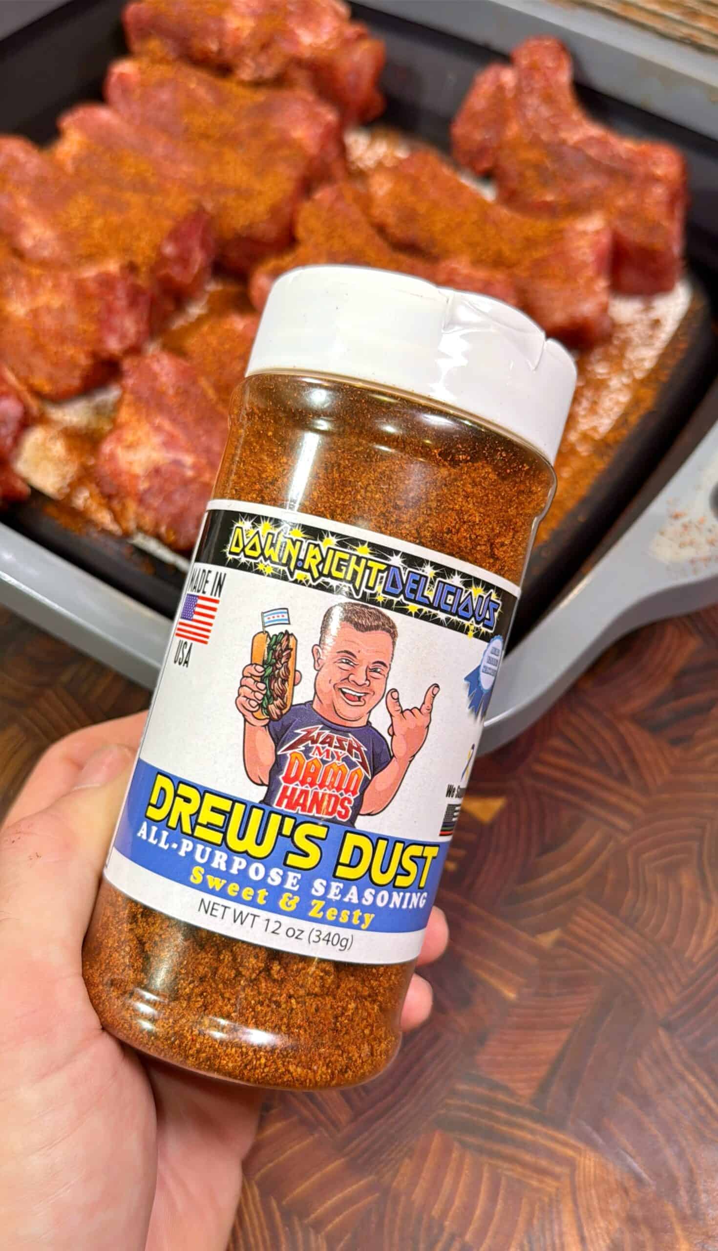 A hand holds a jar of Drew’s Dust All-Purpose Seasoning in front of a tray of raw, seasoned meat. The label features a cartoon man holding the seasoning and reads Down Right Delicious and Sweet & Zesty.