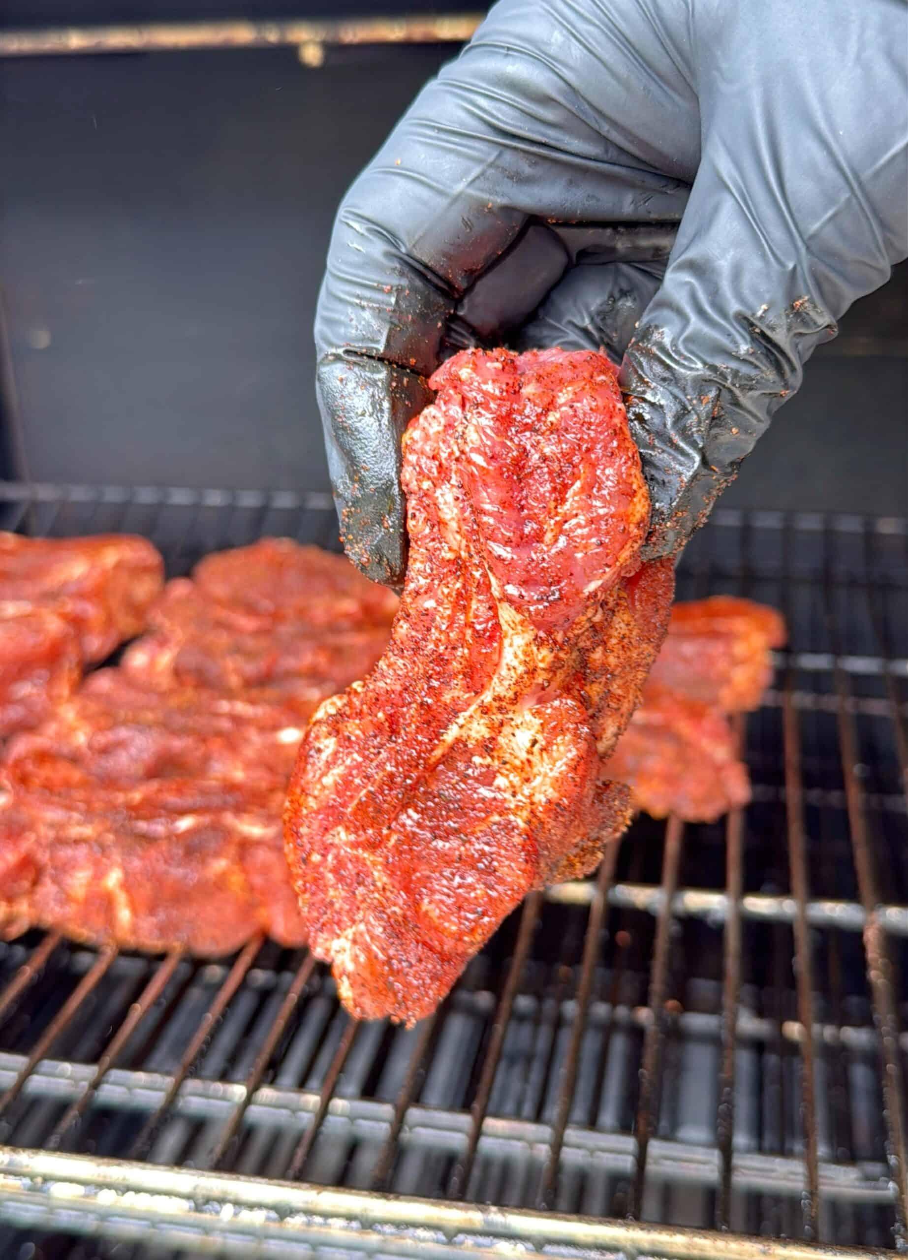A gloved hand holds a seasoned piece of raw meat over a grill with several other pieces of meat cooking in the background.