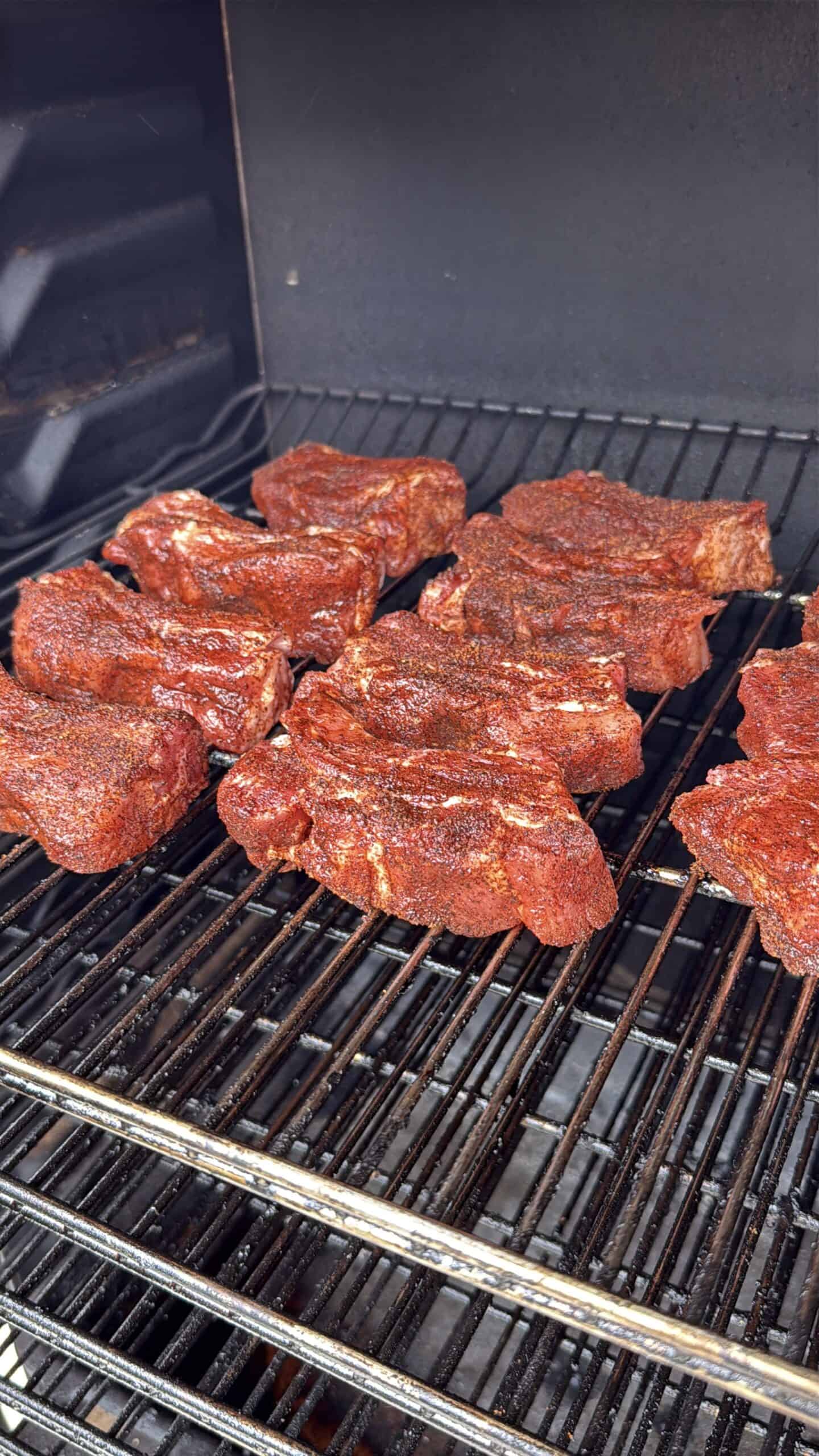 Several pieces of seasoned meat are arranged on the racks inside a smoker or grill, cooking over indirect heat. The meat has a reddish-brown spice rub and is beginning to cook.