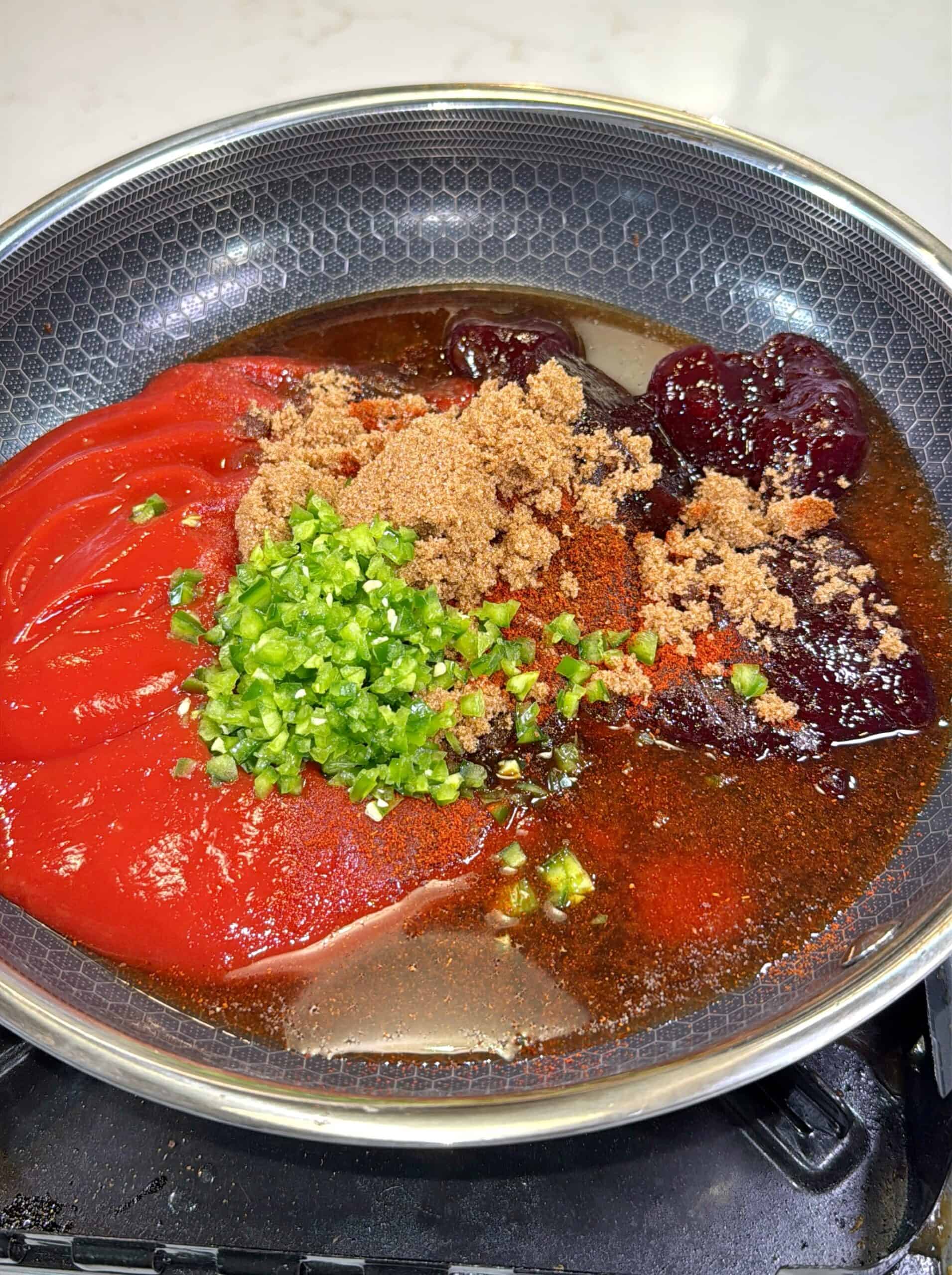 A saucepan on a stovetop filled with ketchup, brown sugar, chopped green peppers, grape jelly, and liquid seasonings, all unmixed, ready to be cooked.