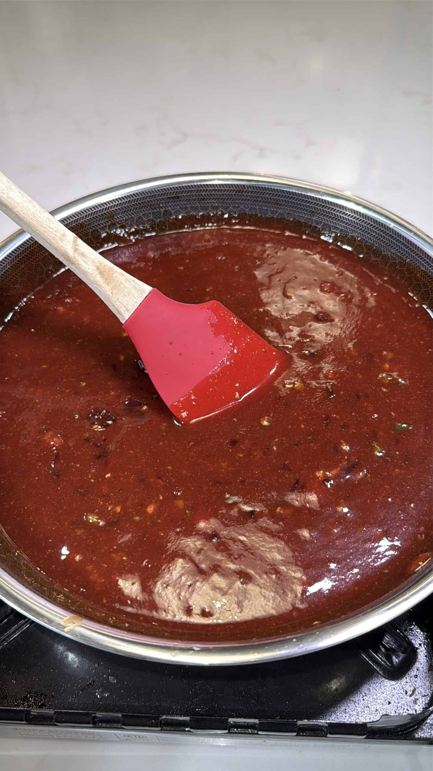 A red silicone spatula stirs a thick, dark red sauce with visible spices in a metal pan on a stovetop. The background is a white countertop.
