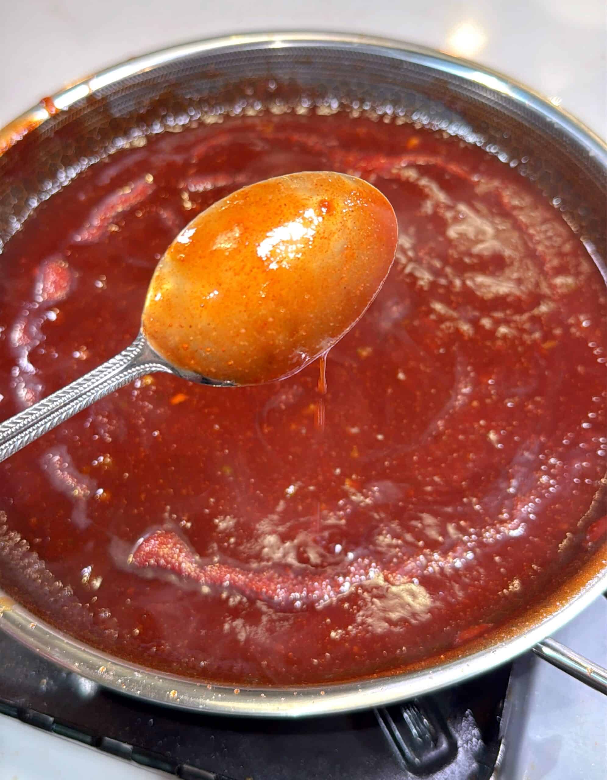 A close-up of a spoon holding thick, red tomato sauce above a simmering pot of sauce on a stovetop. The sauce is glossy and smooth, with visible steam rising.
