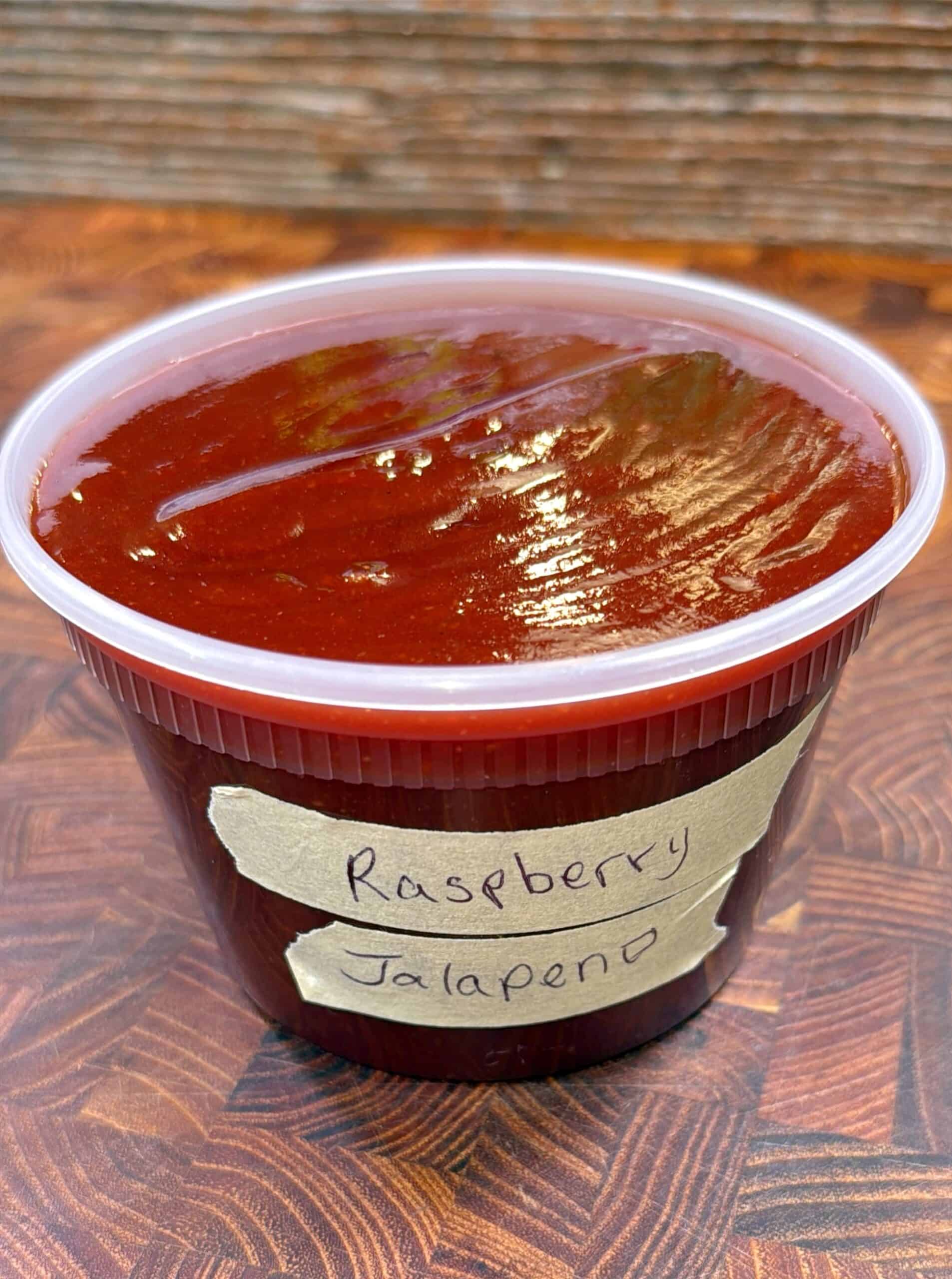 A plastic container filled with deep red sauce sits on a wooden surface. A handwritten label on the container reads Raspberry Jalapeno.