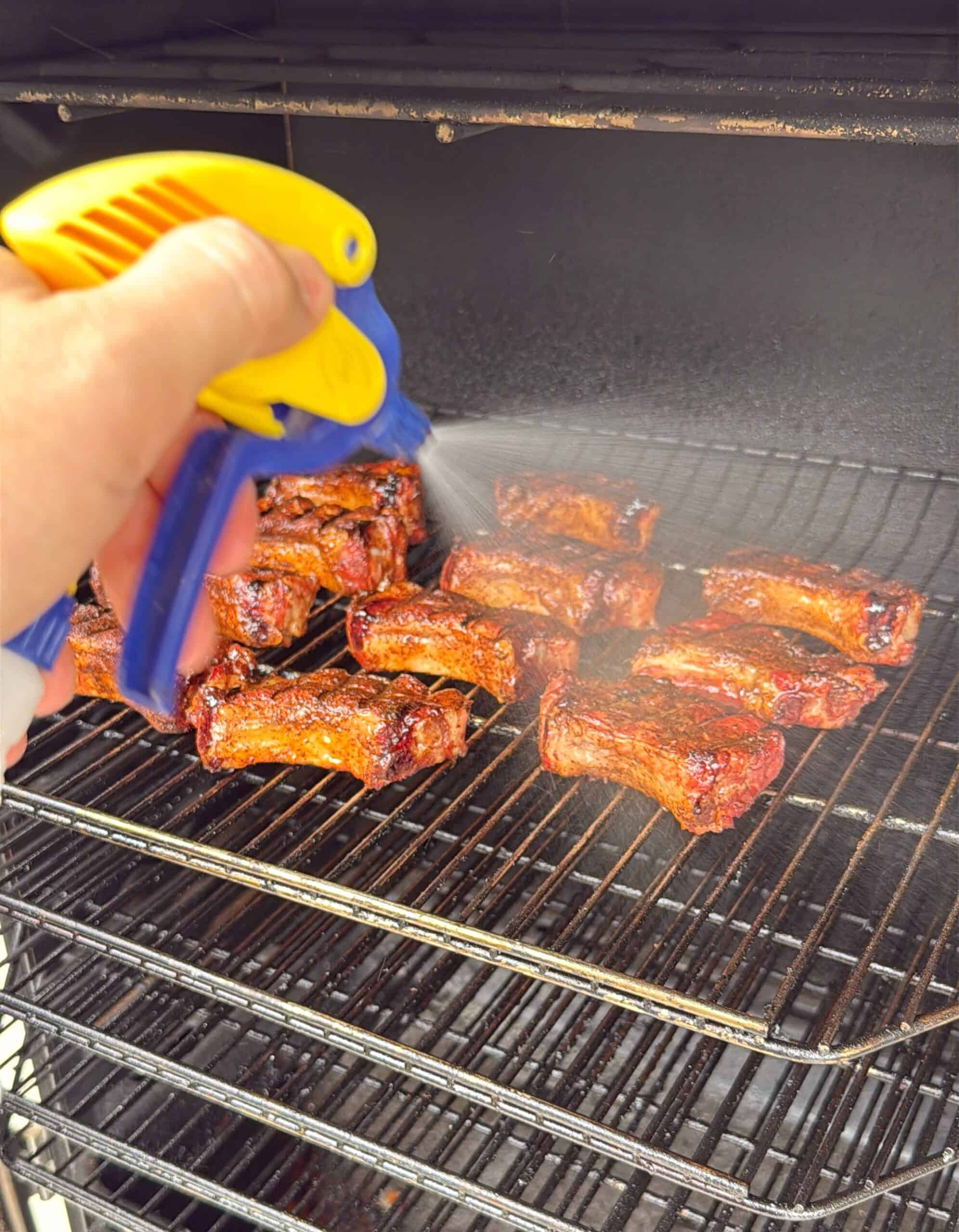 A hand uses a yellow and blue spray bottle to mist several pieces of barbecued meat cooking on the racks of a smoker or grill.