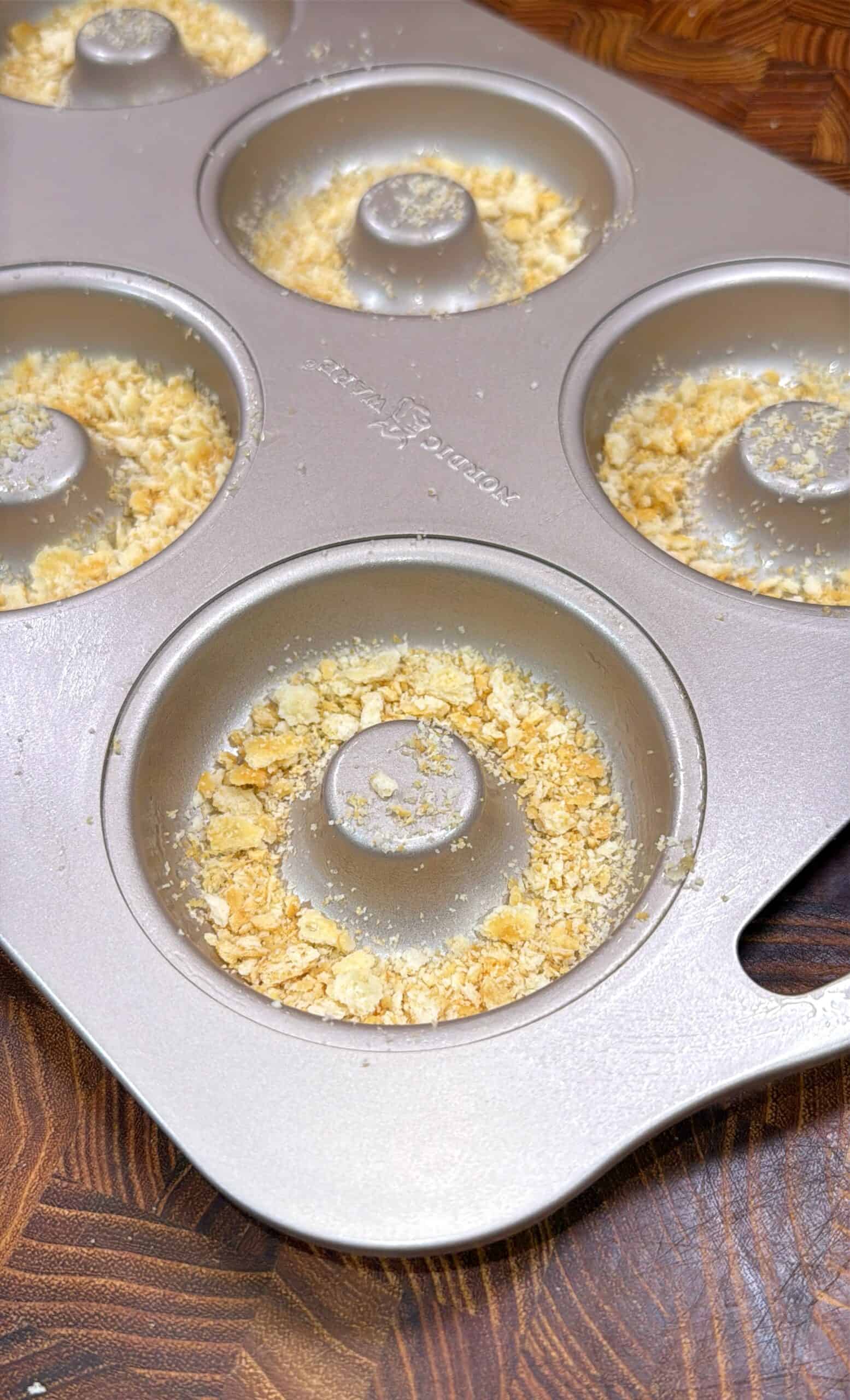 A metal donut baking pan contains a crumbly mixture pressed into the bottom of each mold, set on a wooden surface.