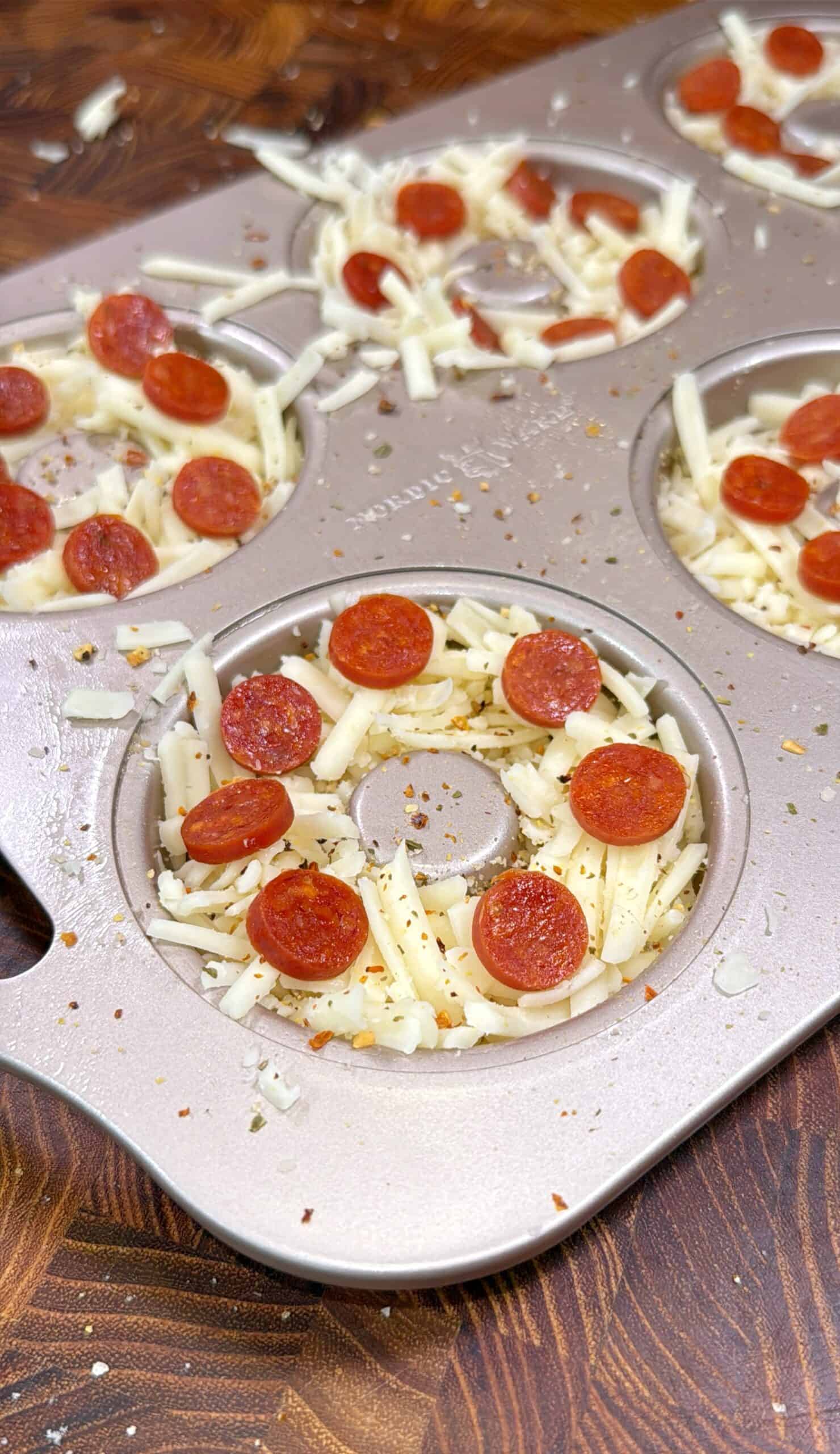 A muffin tin is filled with shredded cheese and mini pepperoni slices, resembling pizza cupcakes, on a wooden surface with scattered herbs and cheese.