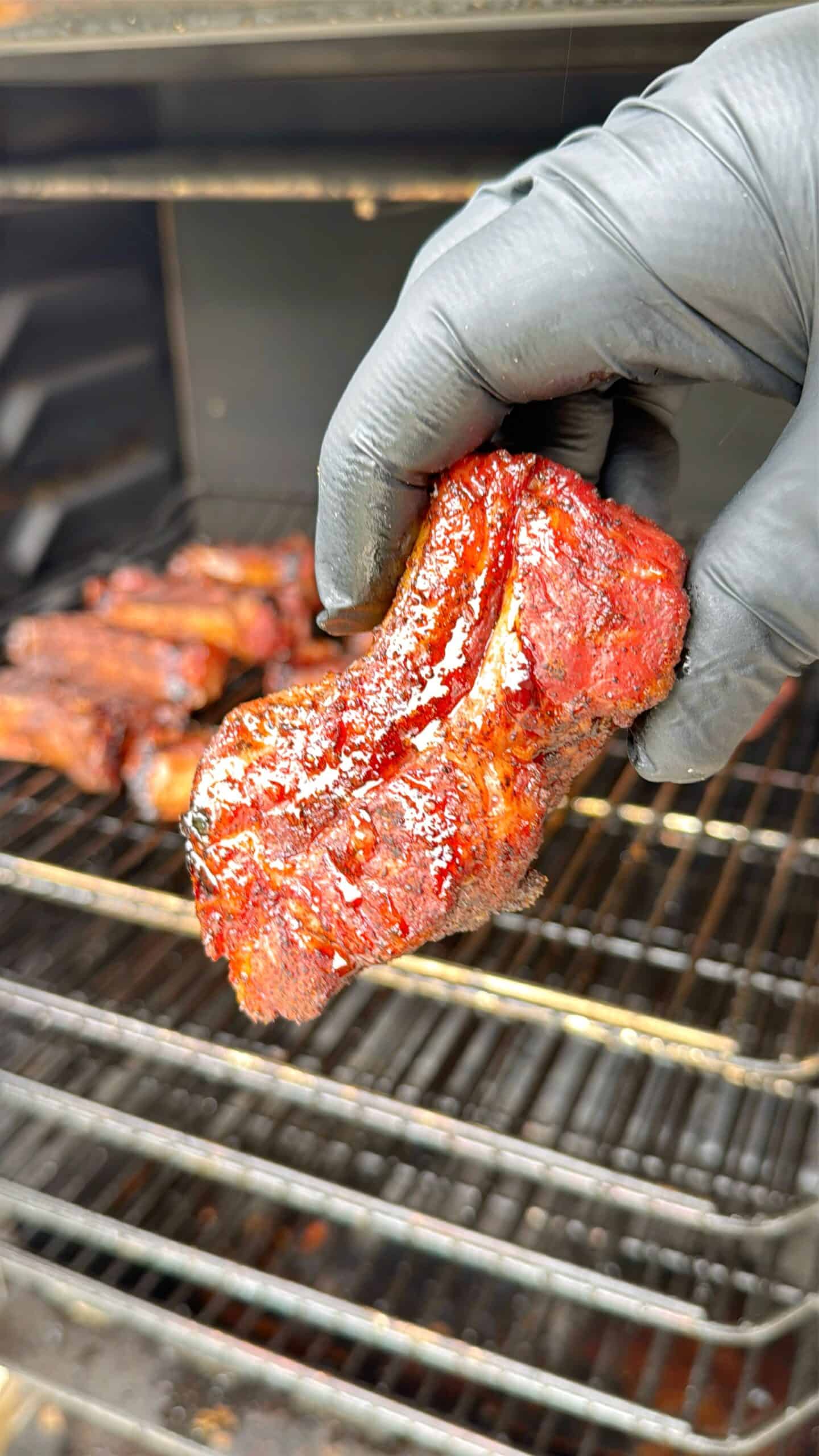 A gloved hand holds a piece of smoky, glazed barbecued meat above an open grill with more pieces of meat cooking on the racks in the background.