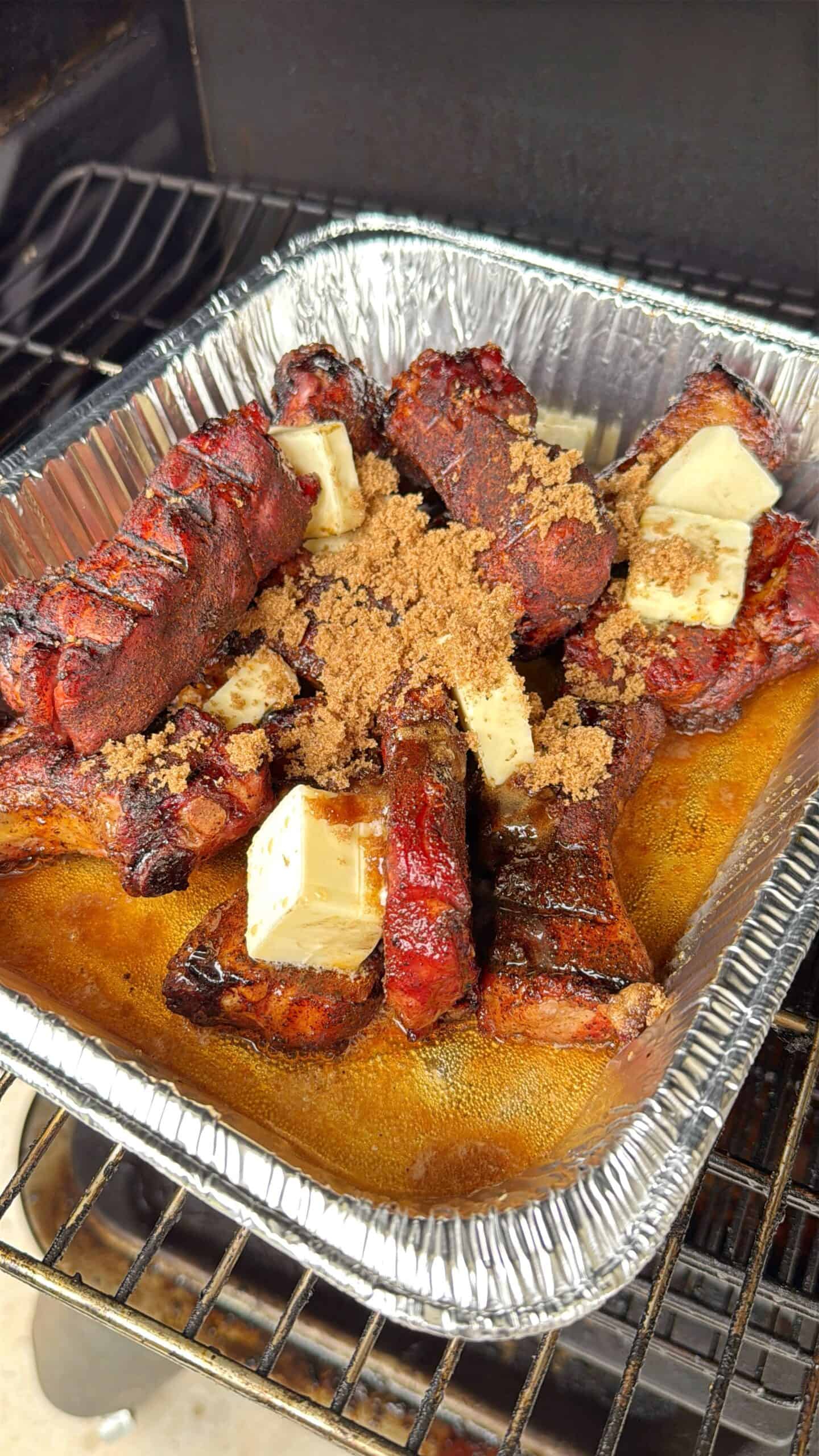 A foil tray filled with smoked pork ribs topped with chunks of butter and sprinkled with brown sugar sits on a barbecue grill. The ribs are coated with a dark, caramelized seasoning.
