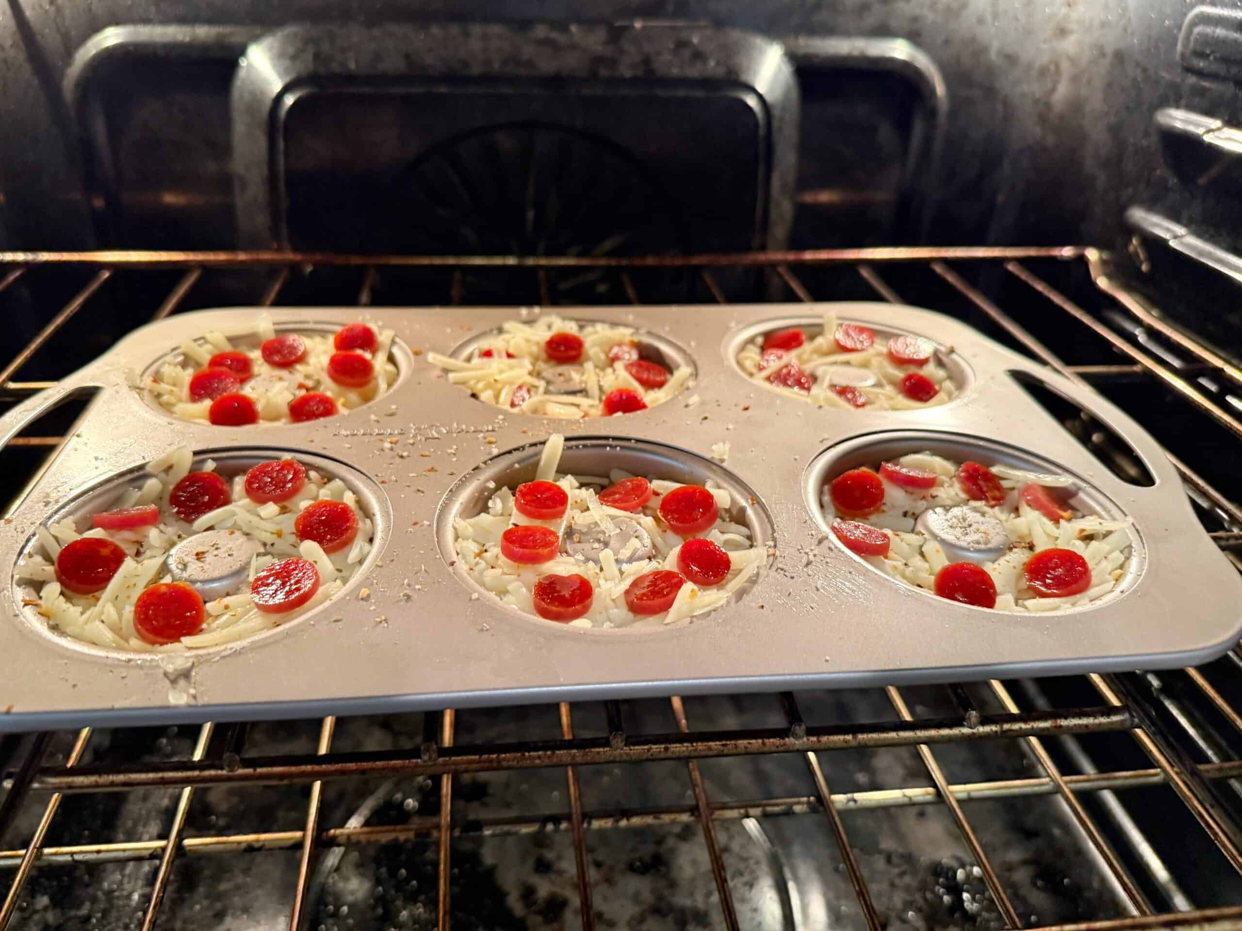 A muffin tin in an oven holds six unbaked mini pizzas topped with cheese, banana slices, and halved cherry tomatoes. The oven racks and interior are visible in the background.