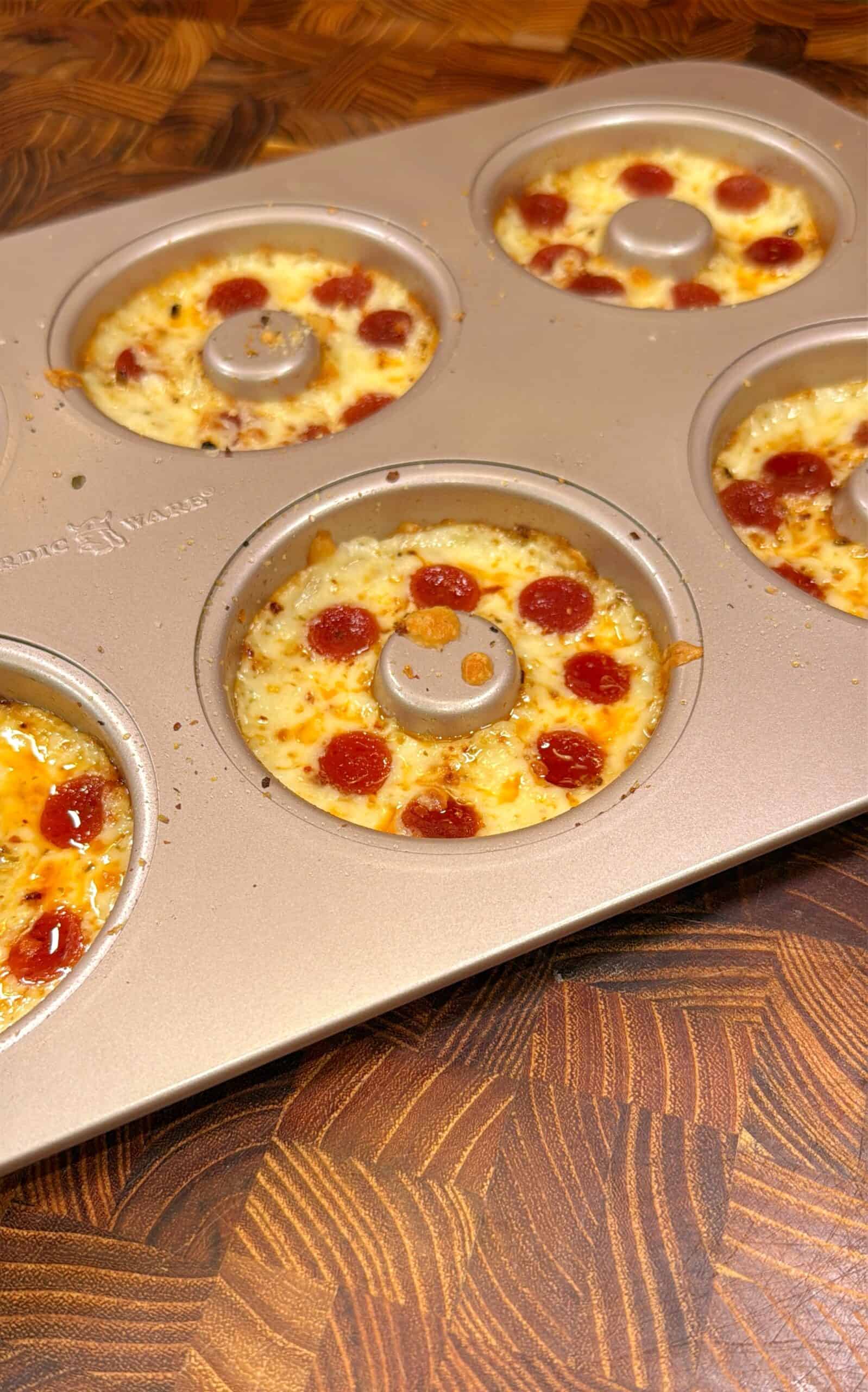 A donut-shaped baking pan holds several mini pizzas topped with melted cheese and small pepperoni slices, all resting on a wooden surface.