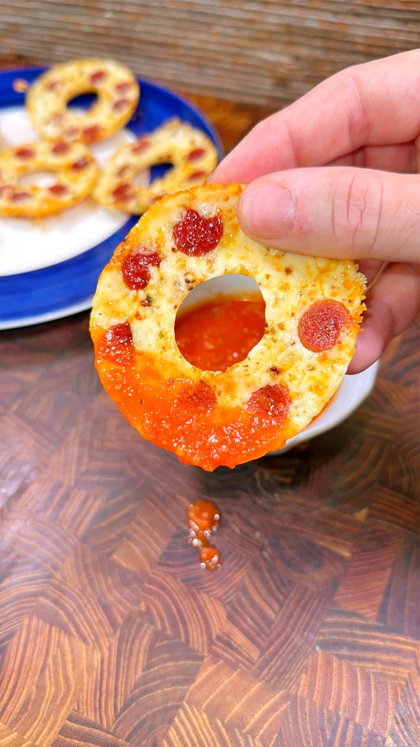 A hand holds a round, hole-in-the-middle mini pizza bagel topped with mini pepperoni, dipping it into a bowl of red sauce, with sauce dripping down. A plate with more bagels is in the background.