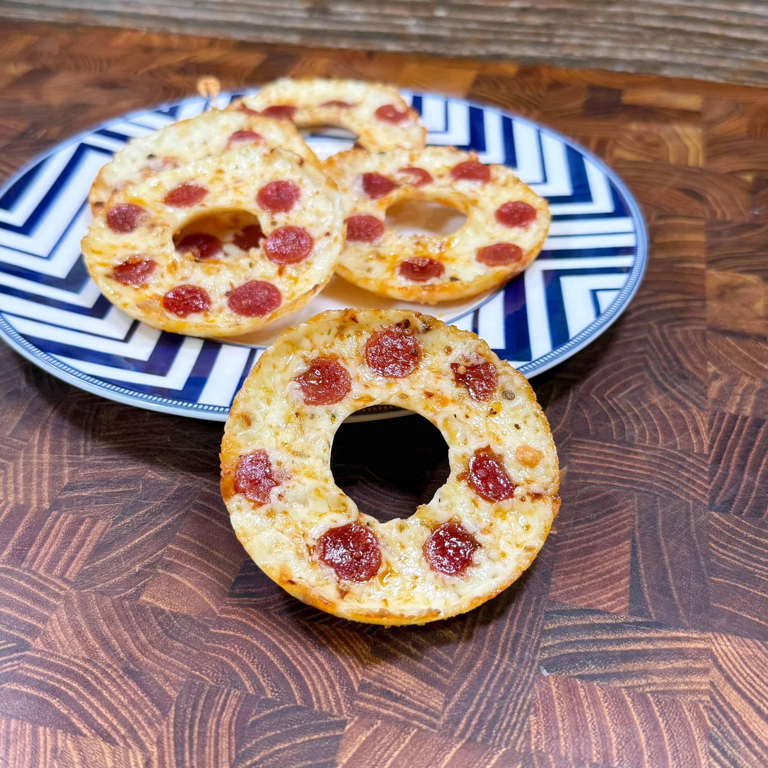 Three bagel halves topped with melted cheese and mini pepperoni are arranged on a blue and white patterned plate, with one bagel half placed in front on a wooden surface.