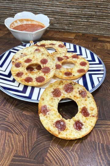 Three bagel halves topped with melted cheese and mini pepperoni slices sit on a patterned plate, with a bowl of marinara sauce for dipping in the background. The setting is a wooden tabletop.