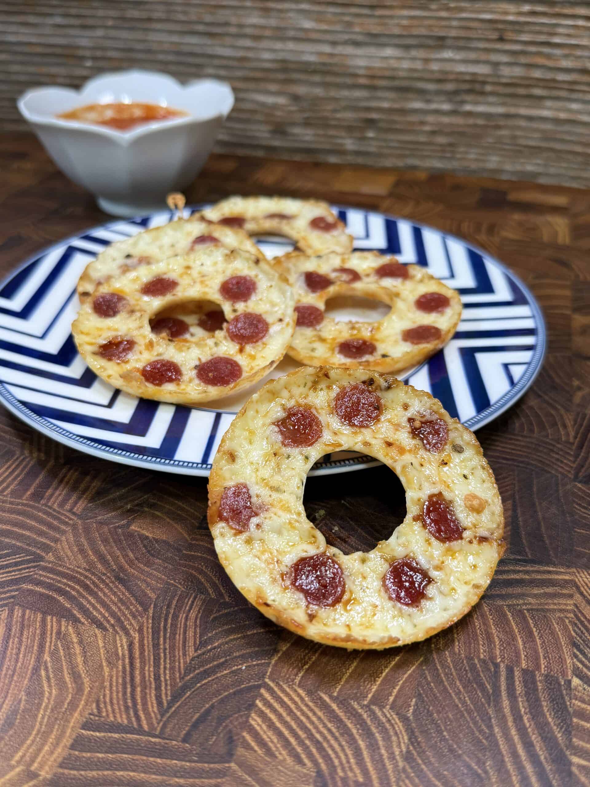 Four bagel-shaped mini pizzas with pepperoni and cheese are arranged on a blue and white patterned plate, with a small bowl of dipping sauce in the background on a wooden surface.