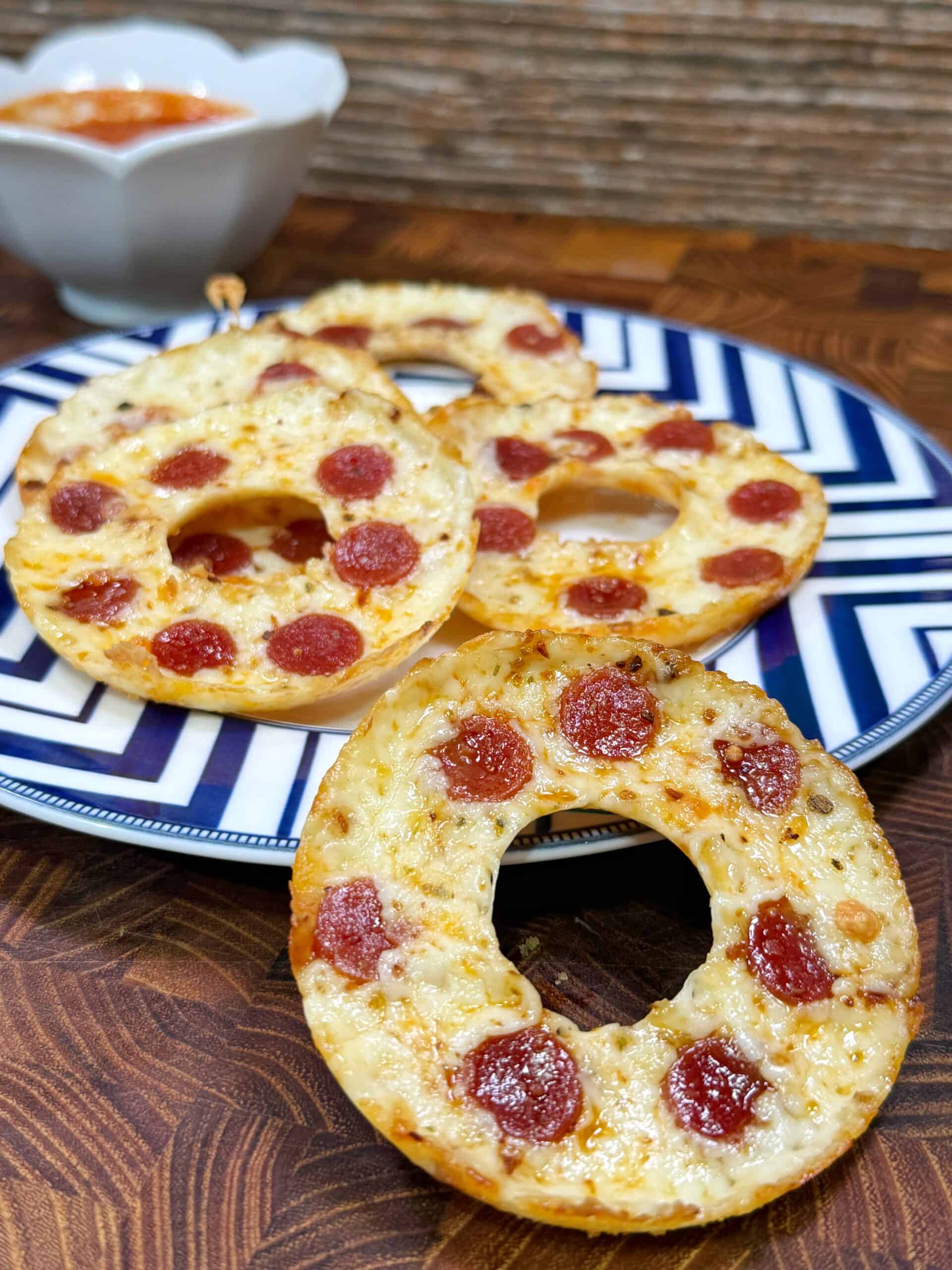 pepperoni pizza rings topped with melted cheese and pepperoni slices are arranged on a plate with a geometric pattern, with a bowl of red dipping sauce in the background on a wooden surface.