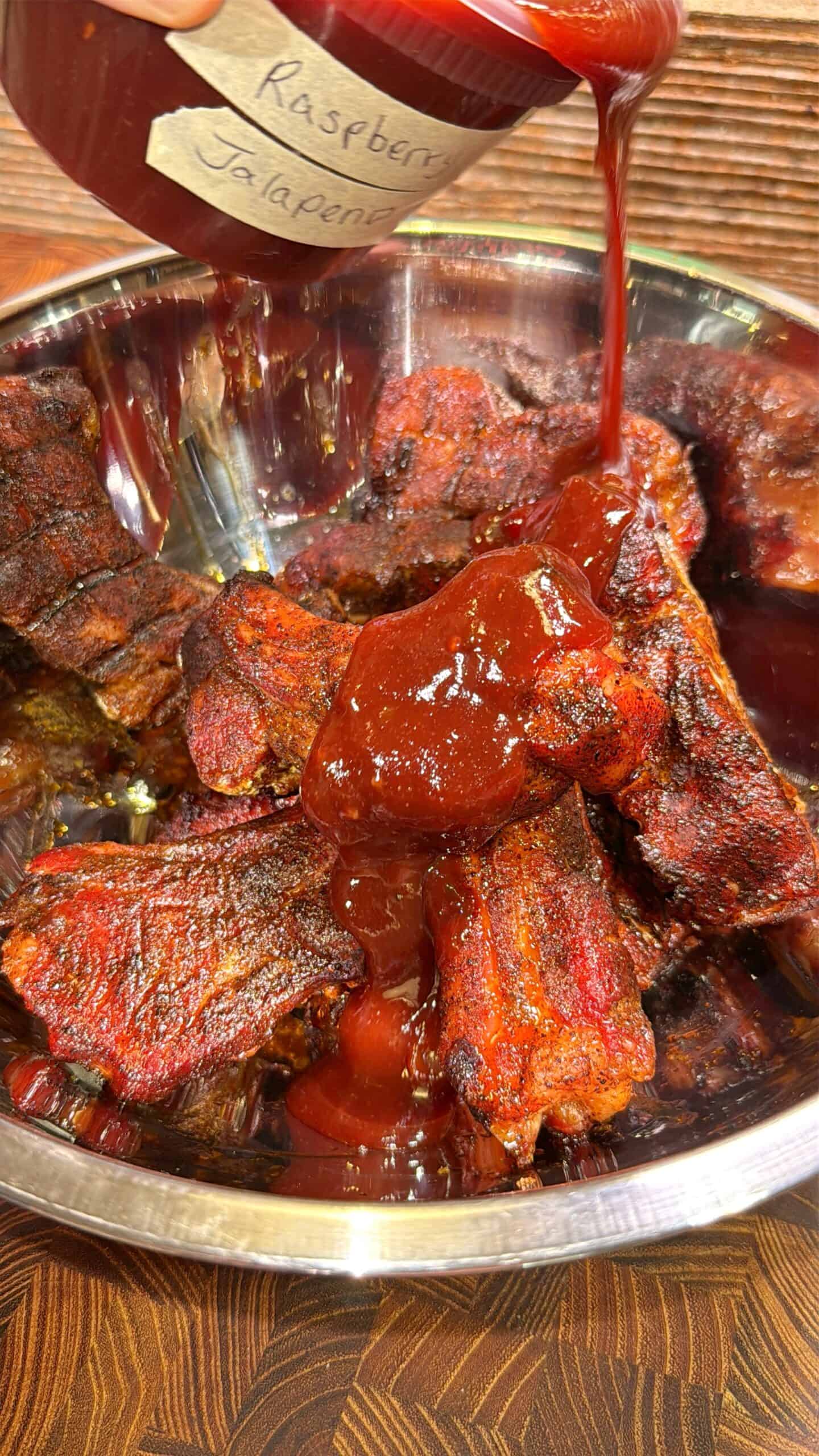 Barbecue ribs in a metal bowl being topped with a thick, red sauce labeled Raspberry Jalapeno from a jar; the sauce is being poured over the ribs.