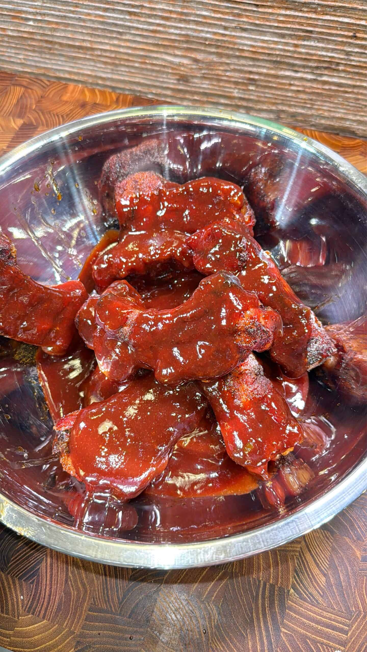 A metal bowl filled with several pieces of chicken coated in a thick, glossy barbecue sauce sits on a wooden surface.